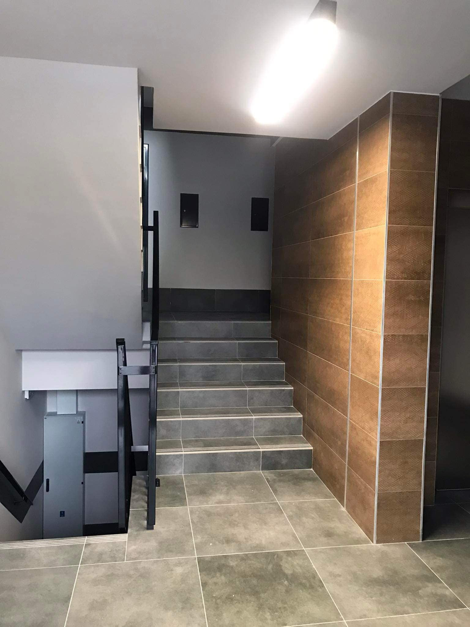 Klatka schodowa w budynku mieszkalnym po remoncie. Szare płytki na schodach i podłodze, brązowe płytki na ścianie, czarna balustrada. Widoczne wejście do windy.