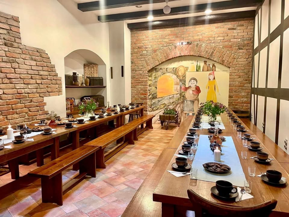 Ustawiony stół w rustykalnej sali z ceglanymi ścianami i malowidłami. Długie drewniane stoły z nakryciami, kwiatami i świecami. Przytulne wnętrze gotowe na przyjęcie.