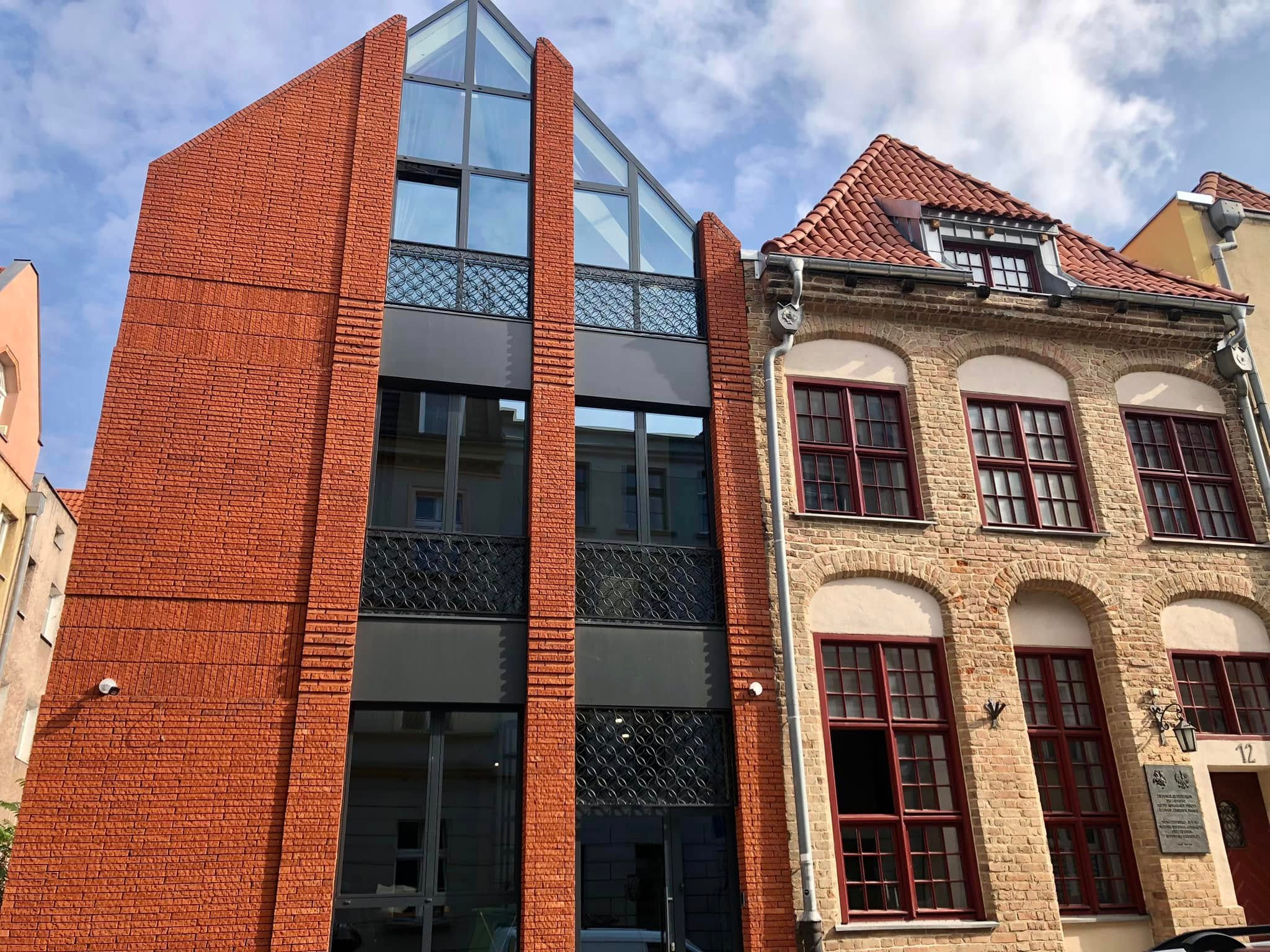 Fasada budynku z czerwonej cegły z nowoczesną, przeszkloną nadbudówką sąsiadująca ze starszą kamienicą z cegły w odcieniach beżu i czerwonymi ramami okiennymi.