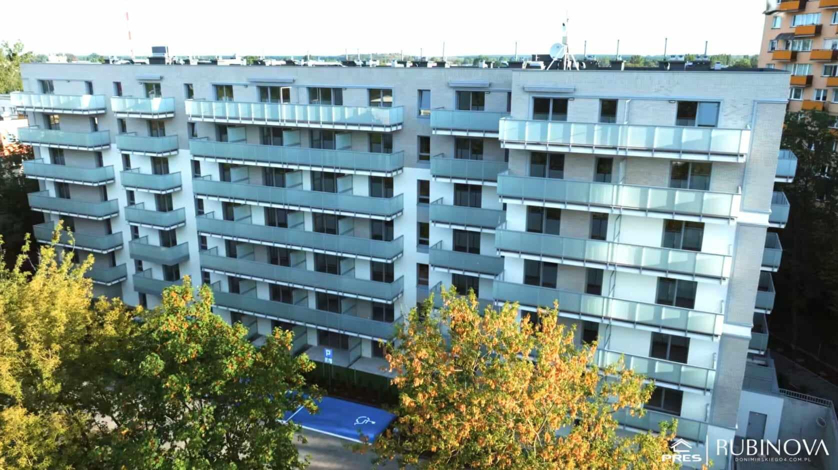 Nowoczesny, wielopiętrowy budynek mieszkalny z balkonami o szklanych balustradach, widziany z perspektywy ptasiej z zielenią drzew na pierwszym planie i miejscem parkingowym.