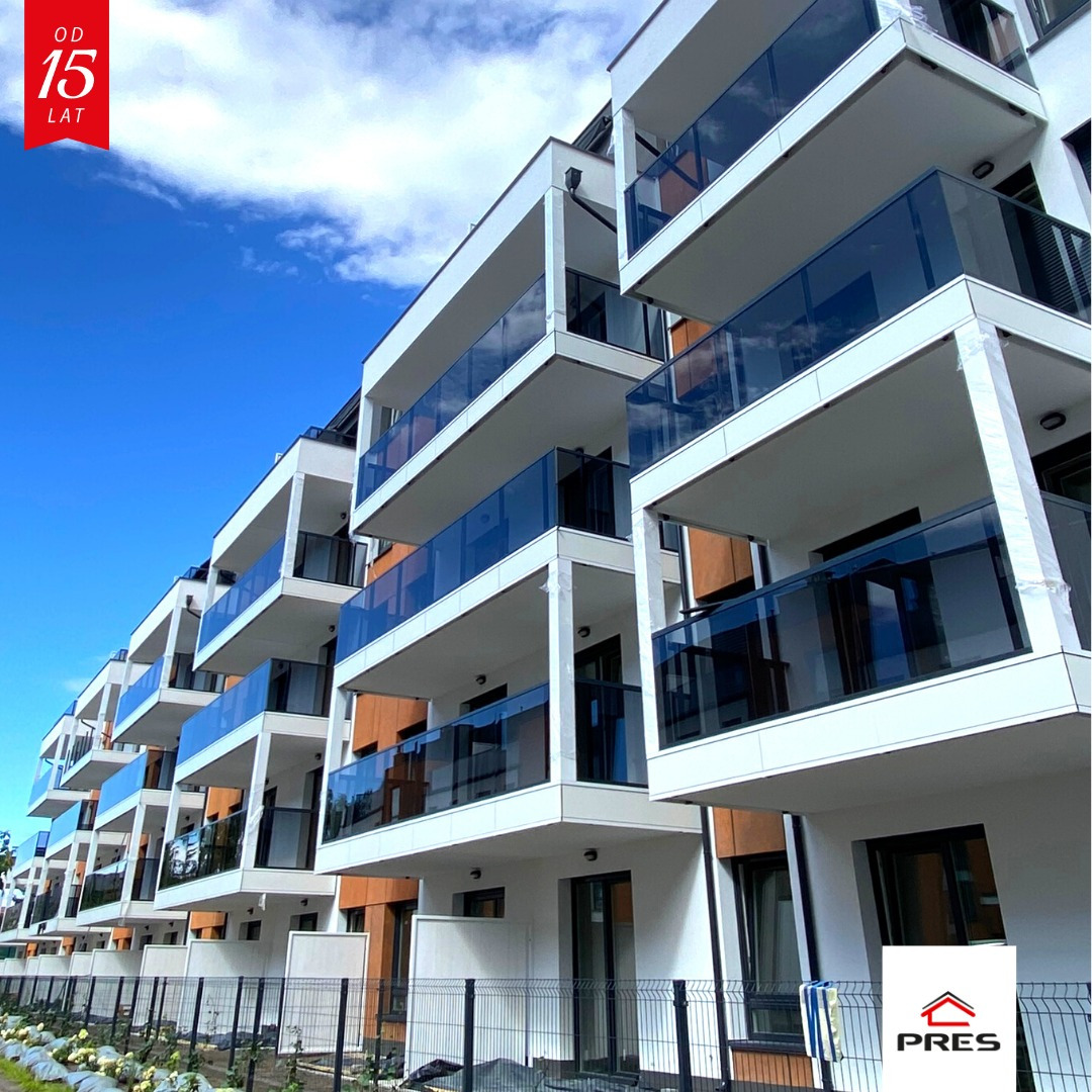 Nowoczesny budynek apartamentowy z białymi balkonami i szklanymi balustradami, widziany pod kątem z dołu, na tle błękitnego nieba. Widoczny fragment ogrodzenia.