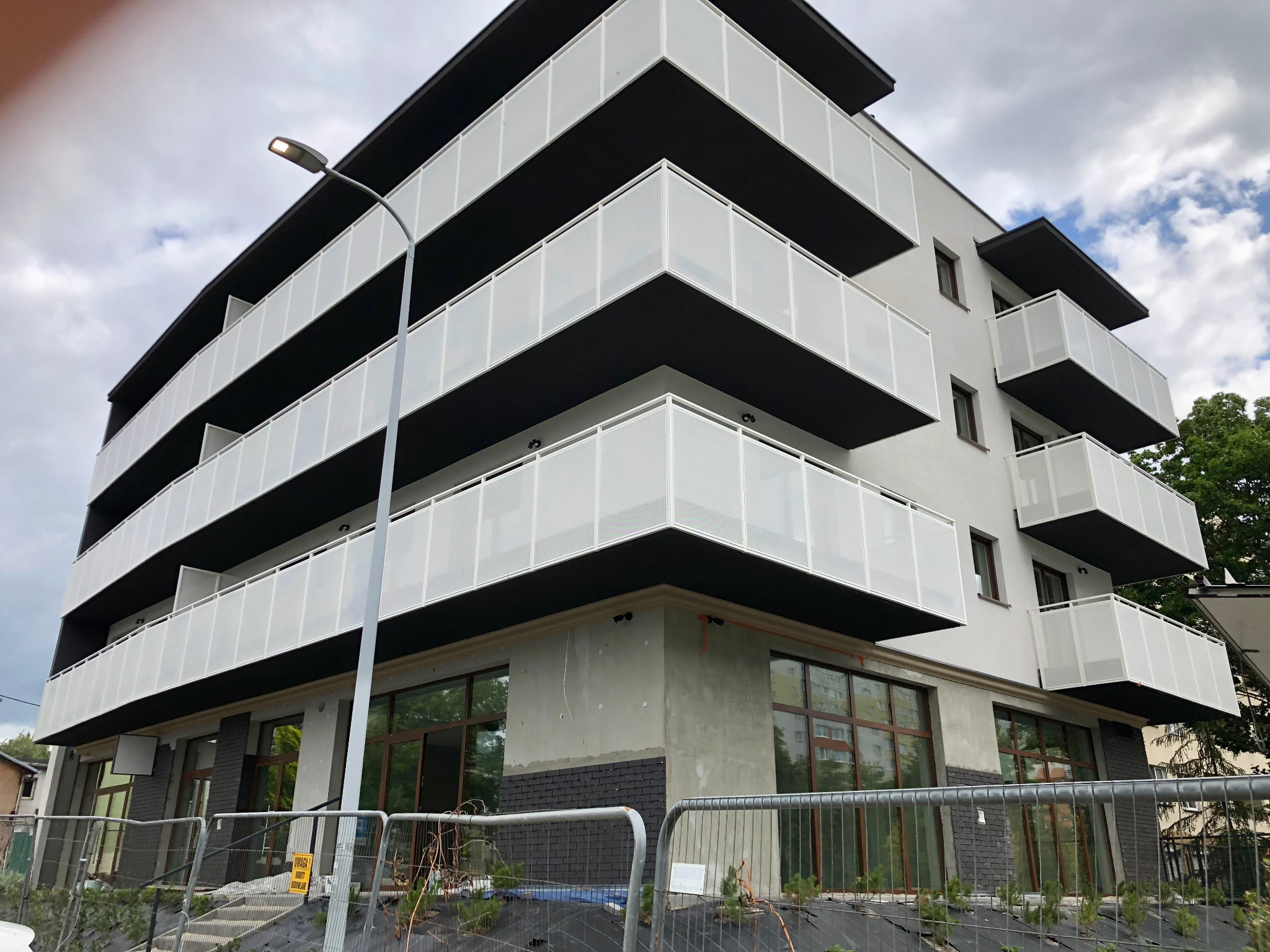 Nowoczesny budynek z balkonami w trakcie wykończenia, widoczne zabezpieczenia budowy i surowy beton na elewacji parteru. Ujęcie z dołu, pochmurne niebo.