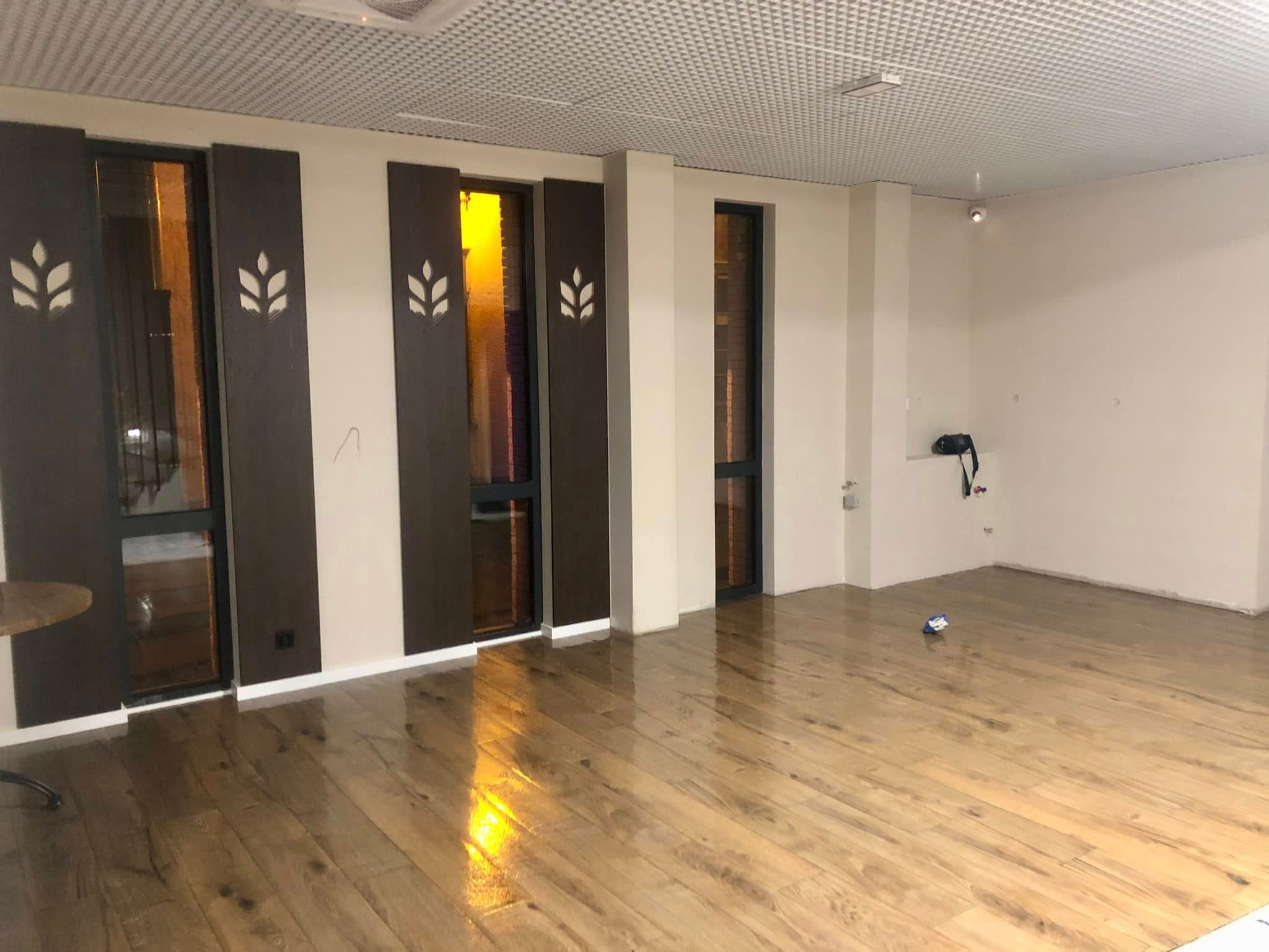 Błyszcząca, drewniana podłoga po umyciu w pustym pomieszczeniu z beżowymi ścianami i ciemnymi drzwiami z dekoracyjnymi panelami. Widoczne drobne ślady po remoncie.