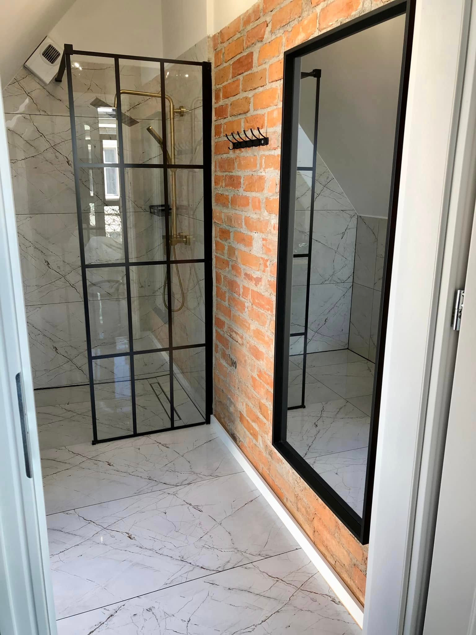 Nowoczesna kabina prysznicowa z czarnymi profilami i złotą armaturą na tle ściany z czerwonej cegły i marmurowych płytek. Widok zza uchylonych drzwi.