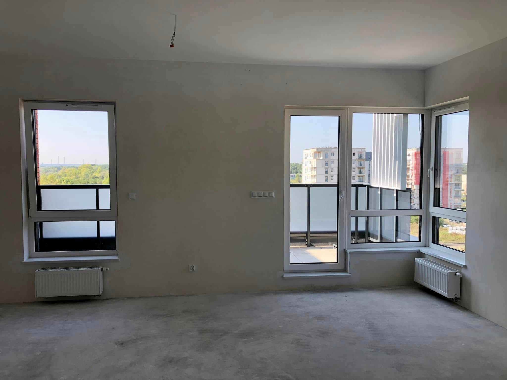 Surowe wnętrze mieszkania po remoncie: gołe ściany, betonowa podłoga, okna z widokiem na zieleń i budynki. Widoczne grzejniki i wystające kable.