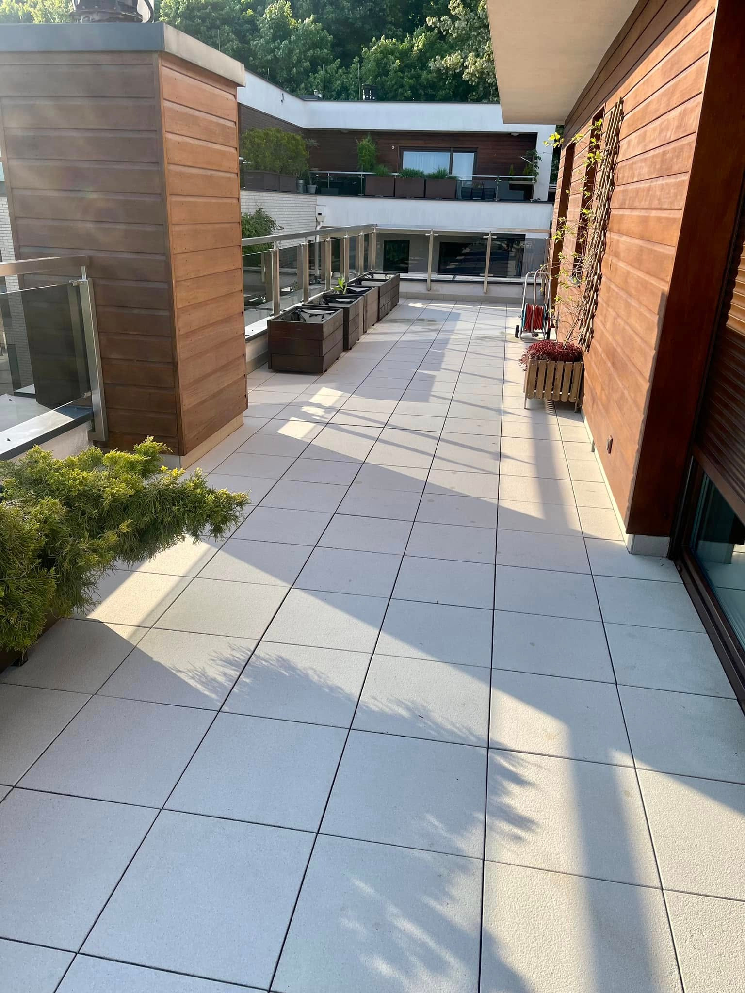 Jasny taras wyłożony kwadratowymi płytkami, z drewnianymi elementami wykończeniowymi i roślinami w donicach. Widoczne refleksy słoneczne i cień drzewa.