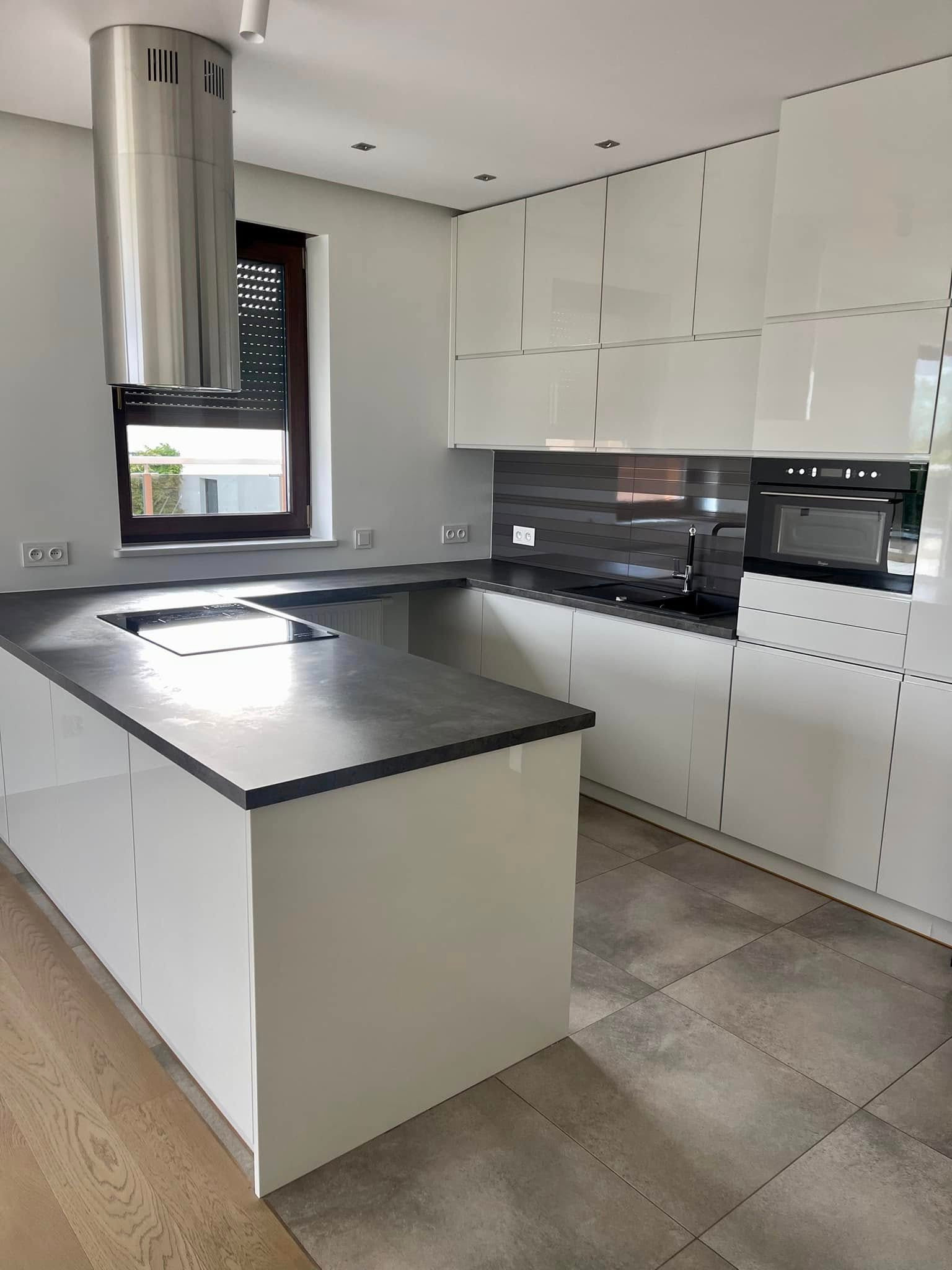 Nowoczesna, jasna kuchnia z wyspą i szarym blatem. Błyszczące białe szafki, stalowy okap cylindryczny i szare płytki nad blatem. Minimalistyczny design.