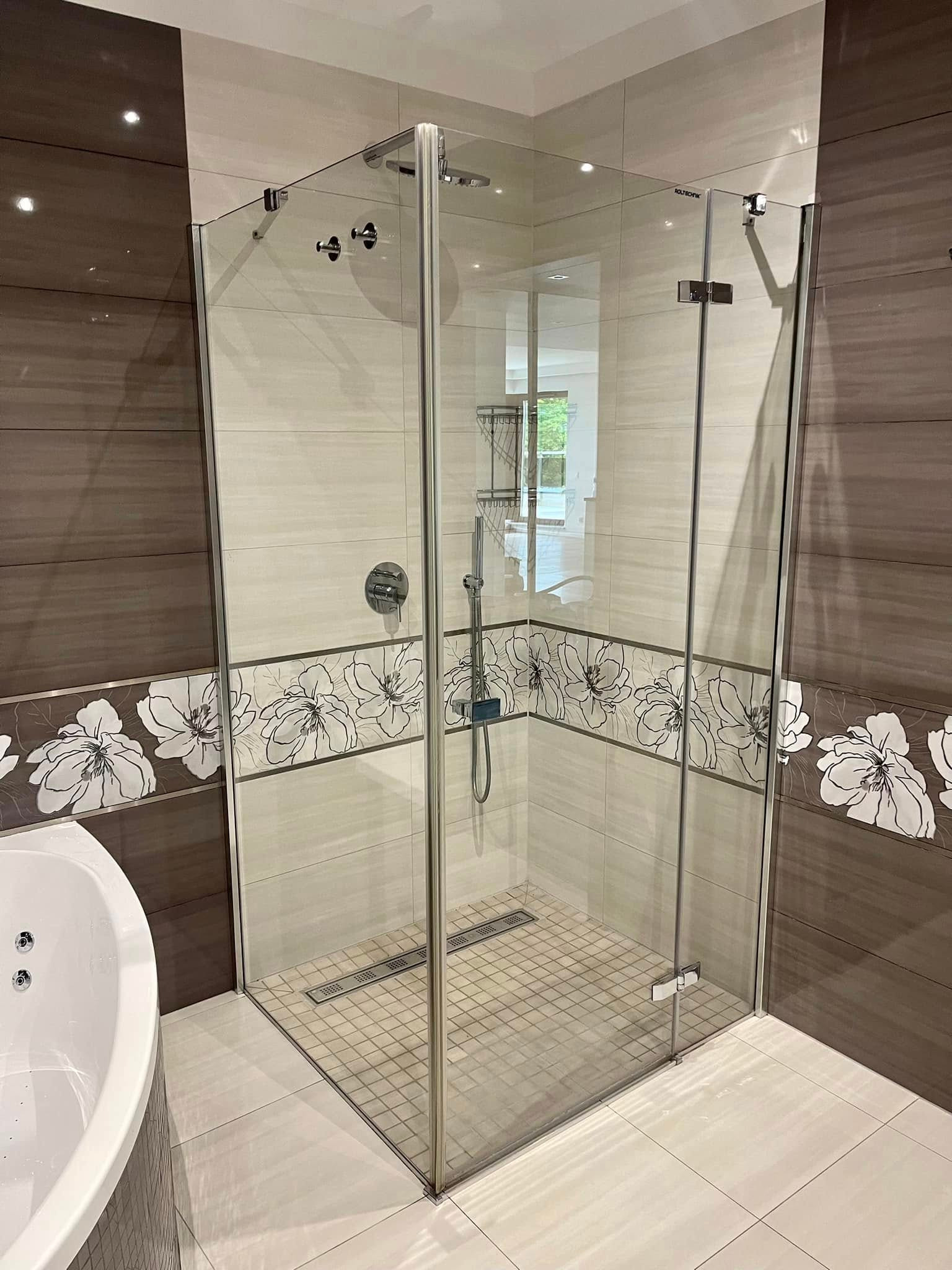 Nowoczesna kabina prysznicowa z chromowanymi okuciami i odpływem liniowym, otoczona beżowo-brązowymi płytkami z motywem kwiatowym. Widoczna część wanny.