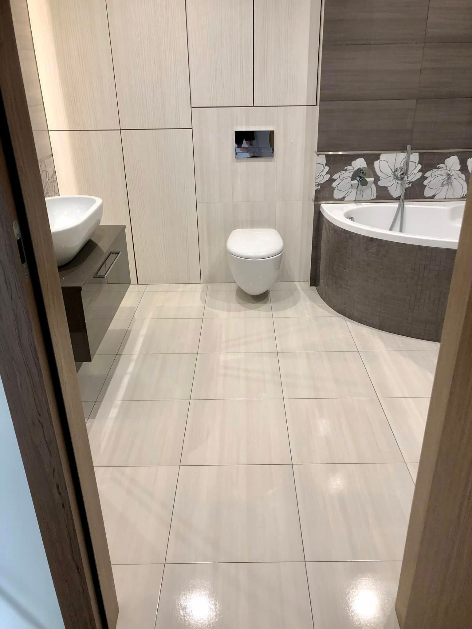 Wykończona łazienka z beżowymi płytkami, białą armaturą i drewnianymi akcentami. Widok zza otwartych drzwi, podkreślający czystość i nowoczesny design.