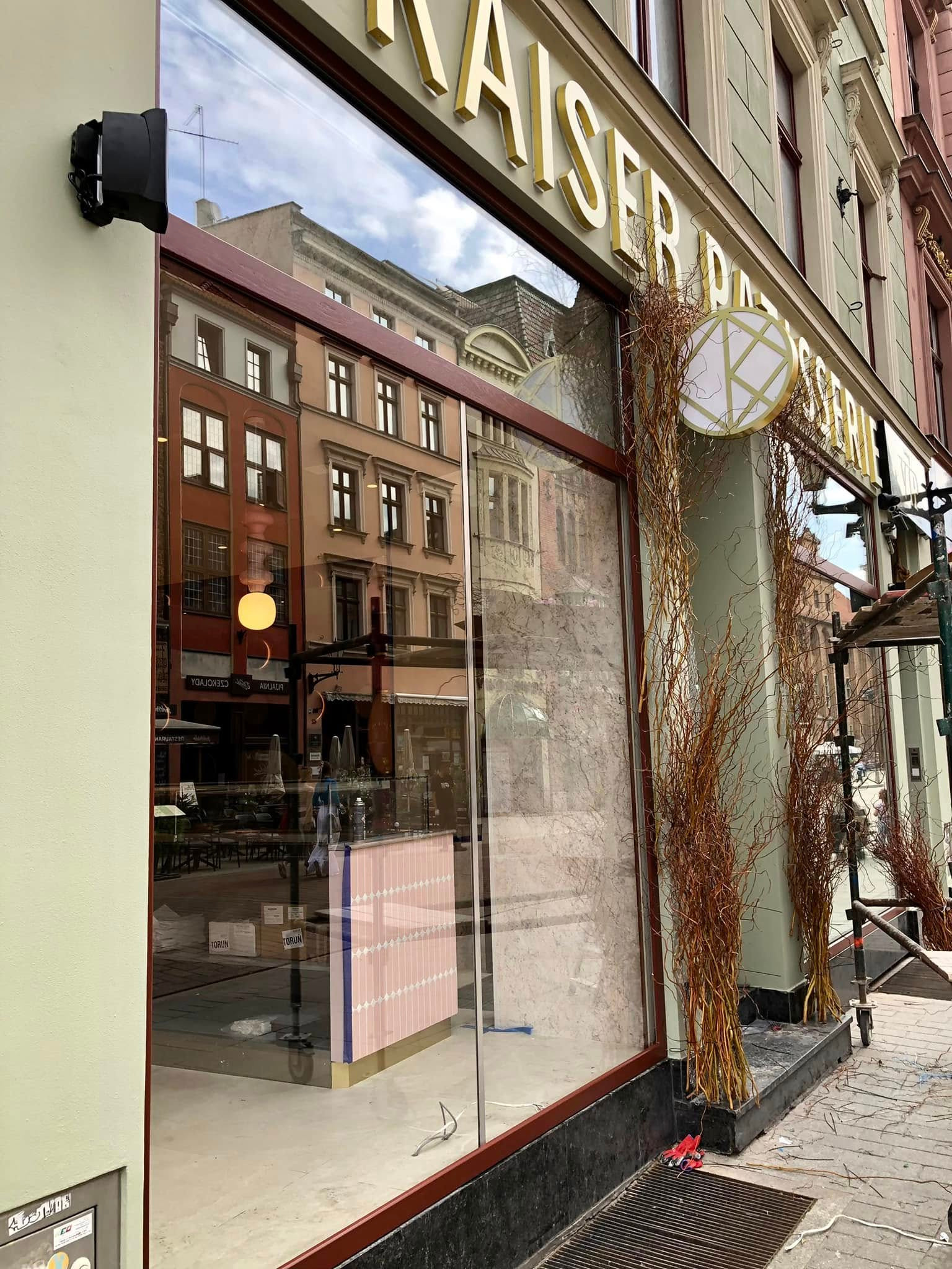Witryna kawiarni 'KAISER PATISSERIE' z dekoracjami z gałązek. W odbiciu widać budynki i wnętrze lokalu. Na chodniku leżą narzędzia.