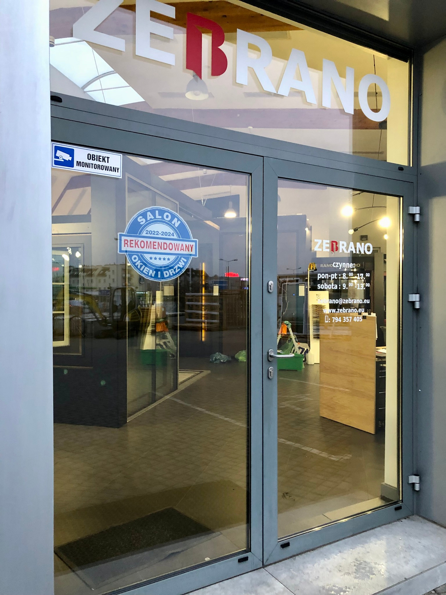 Wejście do salonu okien i drzwi Zebrano, z naklejką 'Salon Rekomendowany 2022-2024' i informacją o monitoringu. Widoczne godziny otwarcia i dane kontaktowe.