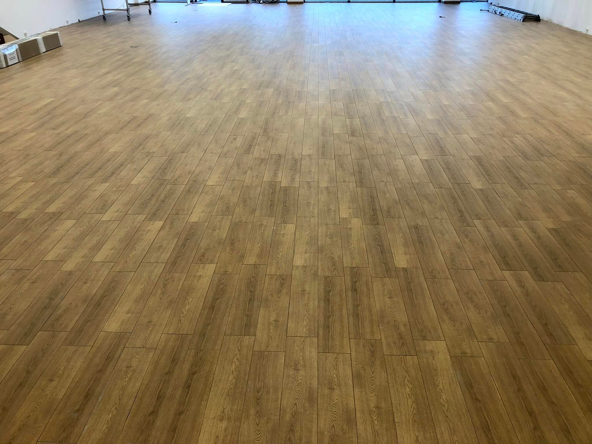 Wnętrze po remoncie: świeżo ułożona, jasnobrązowa podłoga z paneli w dużym, pustym pomieszczeniu. Widoczne kartony i drabina w tle.