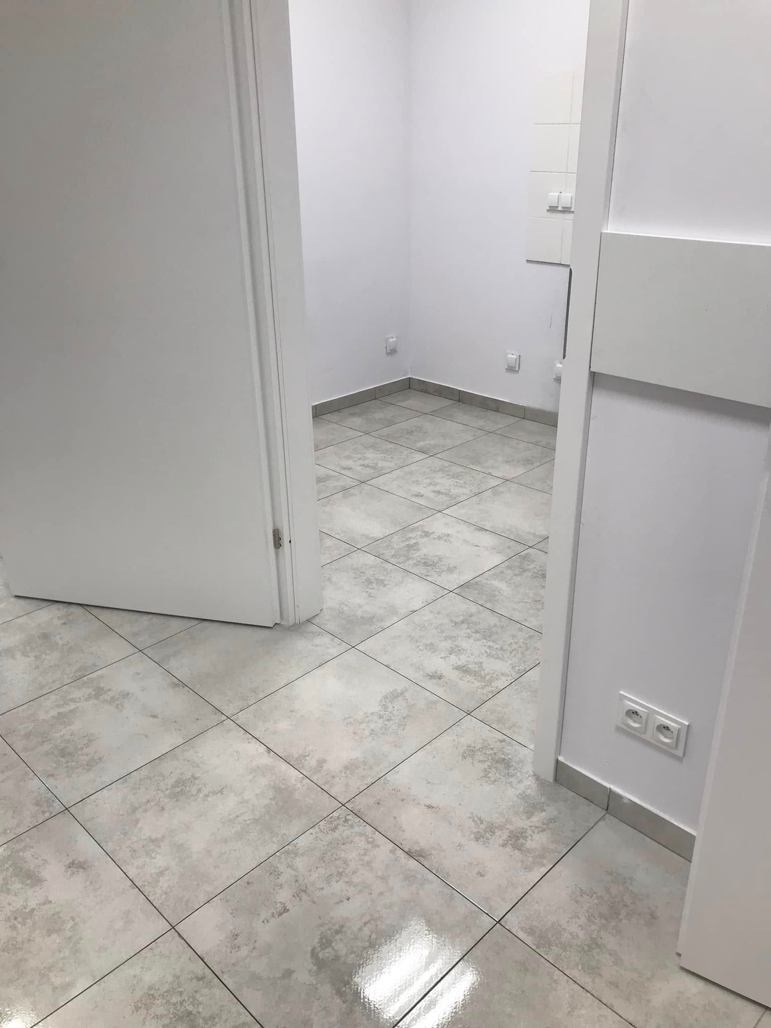 Puste, jasne pomieszczenie z szarą, kwadratową podłogą z płytek, białe ściany i otwarte drzwi. Widoczny fragment gniazdka elektrycznego przy listwie przypodłogowej.