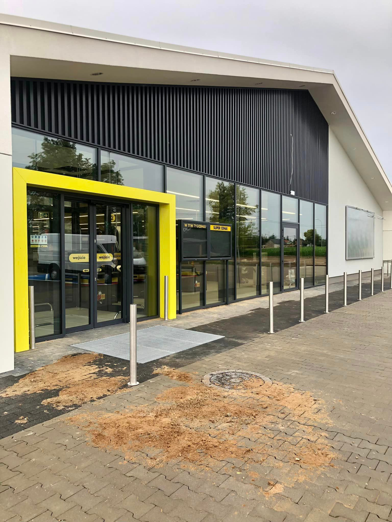 Nowoczesny budynek sklepu z żółtą ramą wejścia i szklaną fasadą. Na kostce brukowej widoczne zabrudzenia ziemią, sugerujące niedawne prace budowlane.