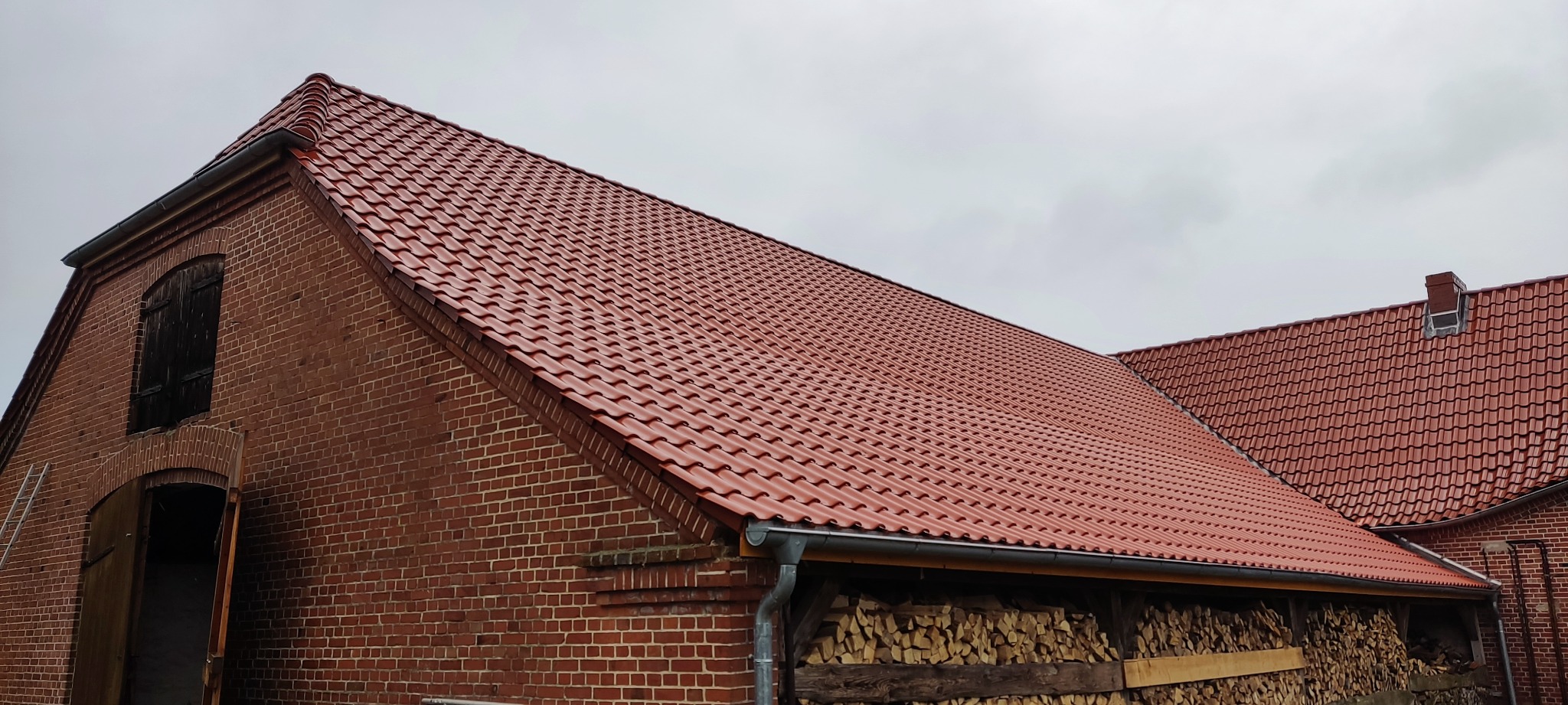 Ceglany budynek z nowym, czerwonym dachem z dachówki ceramicznej. Widoczne rynny i składowane drewno. Pochmurne niebo w tle. Ujęcie z dołu.