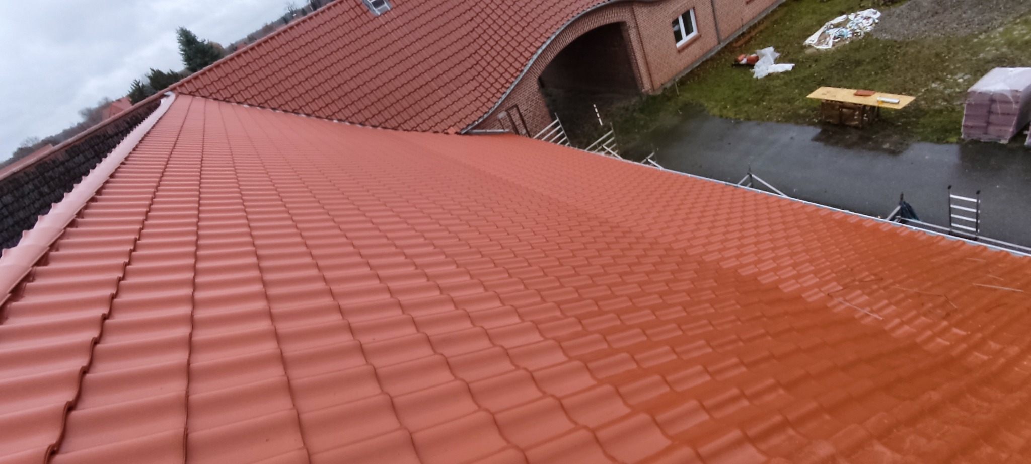 Czerwona dachówka ceramiczna na dachu domu jednorodzinnego, widok z góry, z widocznymi elementami konstrukcyjnymi i otoczeniem budynku. Dach po renowacji.