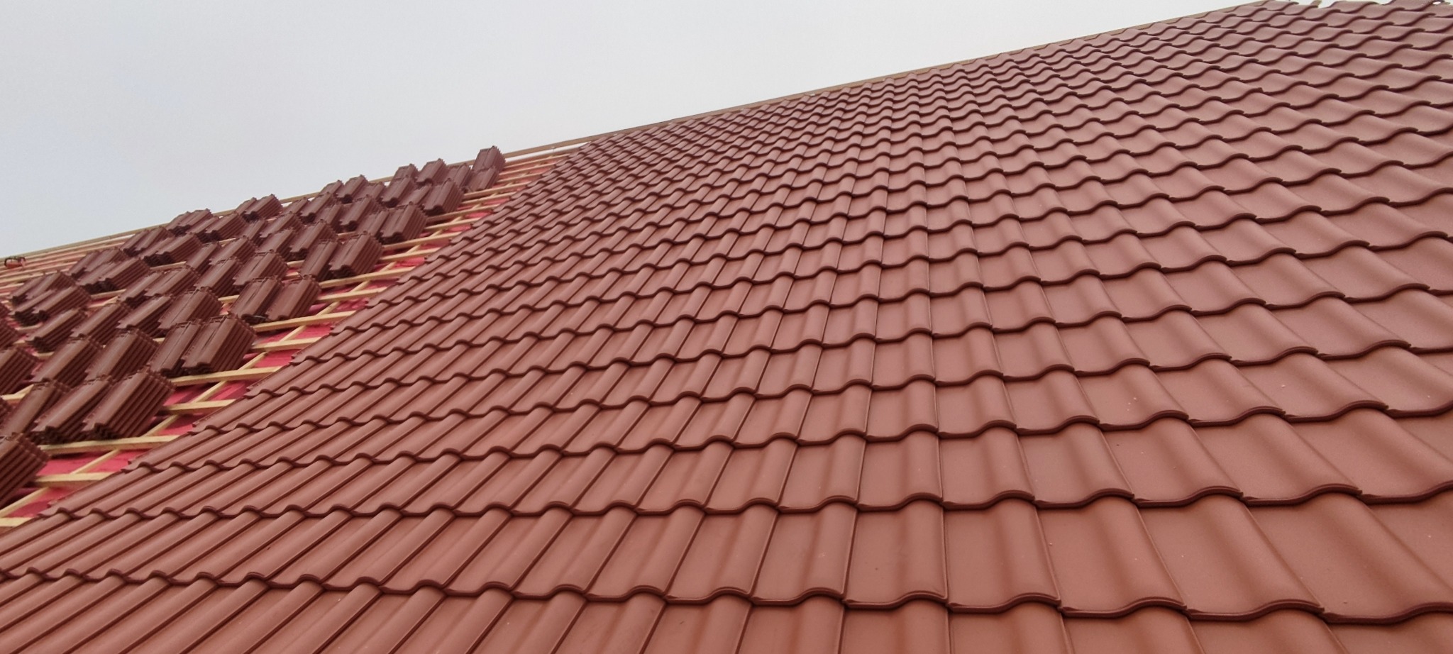 Częściowo pokryty dach z czerwonej blachodachówki, widoczne drewniane elementy konstrukcji i przygotowane do montażu arkusze. Ujęcie z dołu pod kątem.