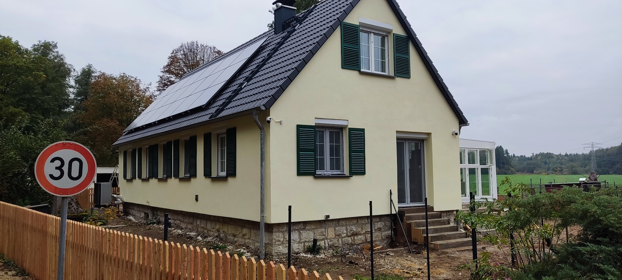 Elewacja domu jednorodzinnego z panelami słonecznymi, zielonymi okiennicami i nowym ogrodzeniem. Widoczny znak drogowy '30'.