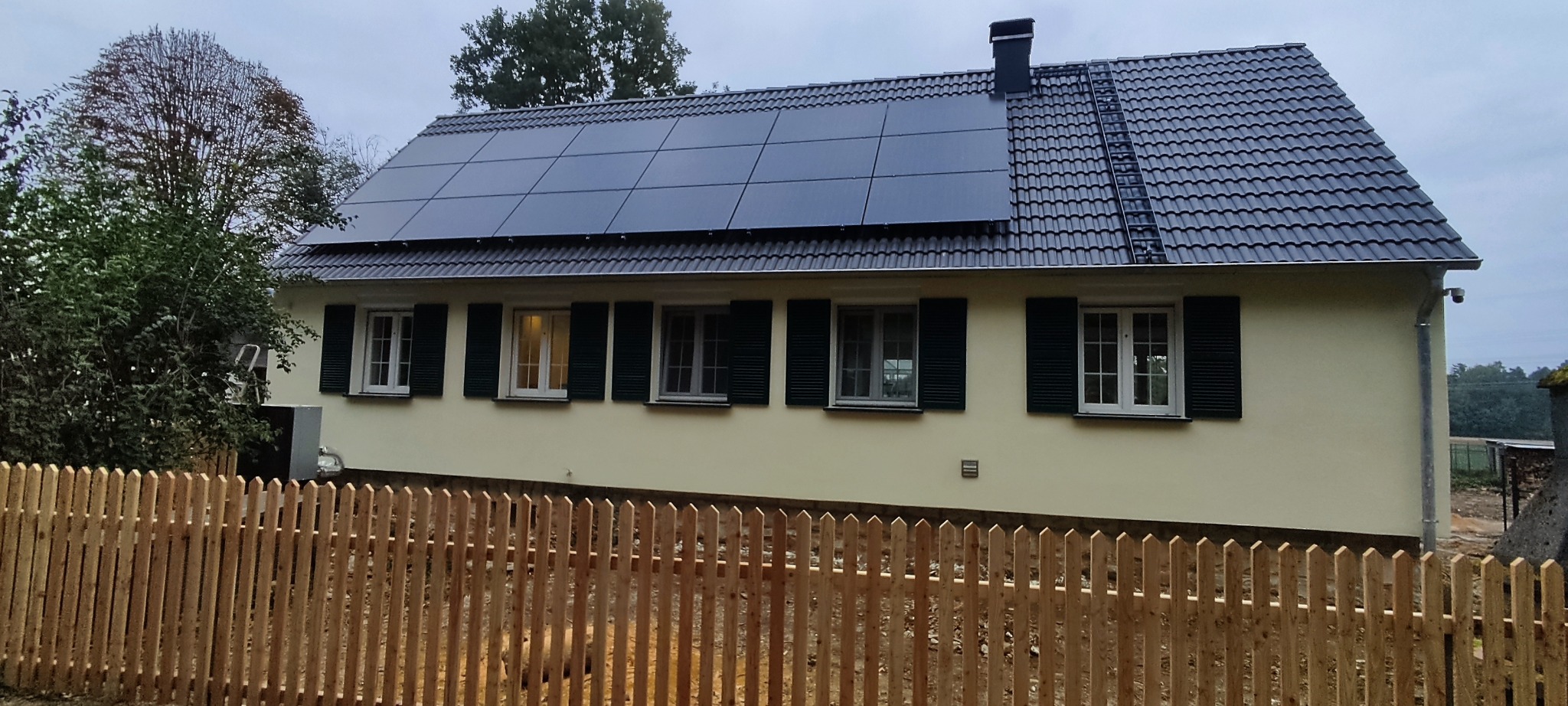 Dom jednorodzinny z panelami słonecznymi na dachu i nowym ogrodzeniem. Elewacja w jasnym odcieniu, okna z zielonymi okiennicami. Widok z poziomu gruntu.