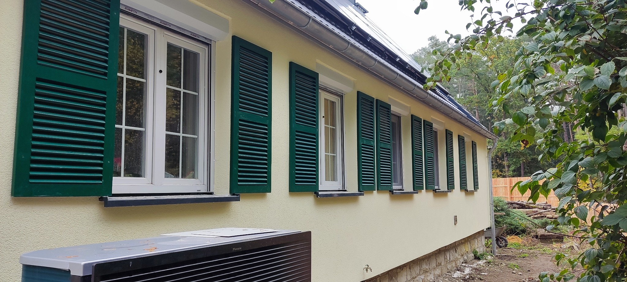 Elewacja domu z kremową fasadą i zielonymi okiennicami. Widoczne białe okna z podziałami i fragment klimatyzatora. W tle drzewa i fragment dachu.