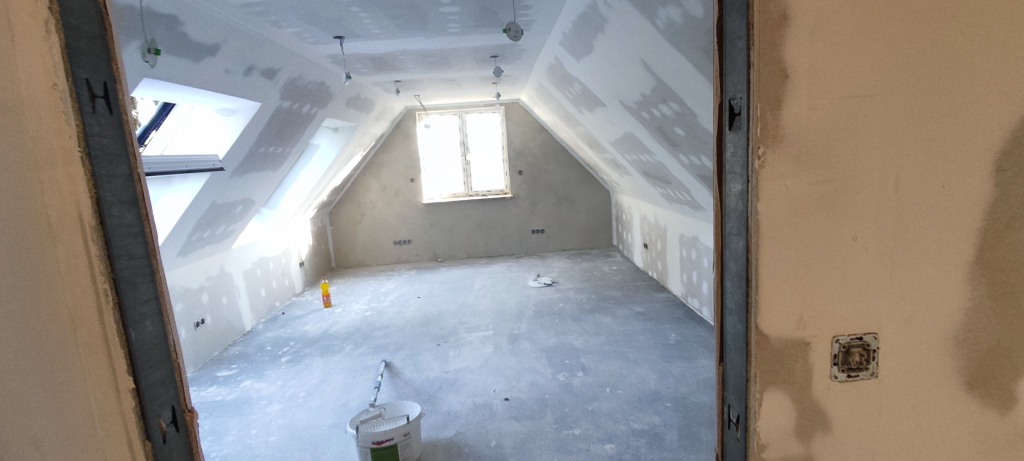 Poddasze w trakcie prac wykończeniowych: szpachlowane ściany i sufit, widoczne okno dachowe, puszki elektryczne, wiadro z farbą i wałek malarski. Ujęcie z perspektywy wejścia.