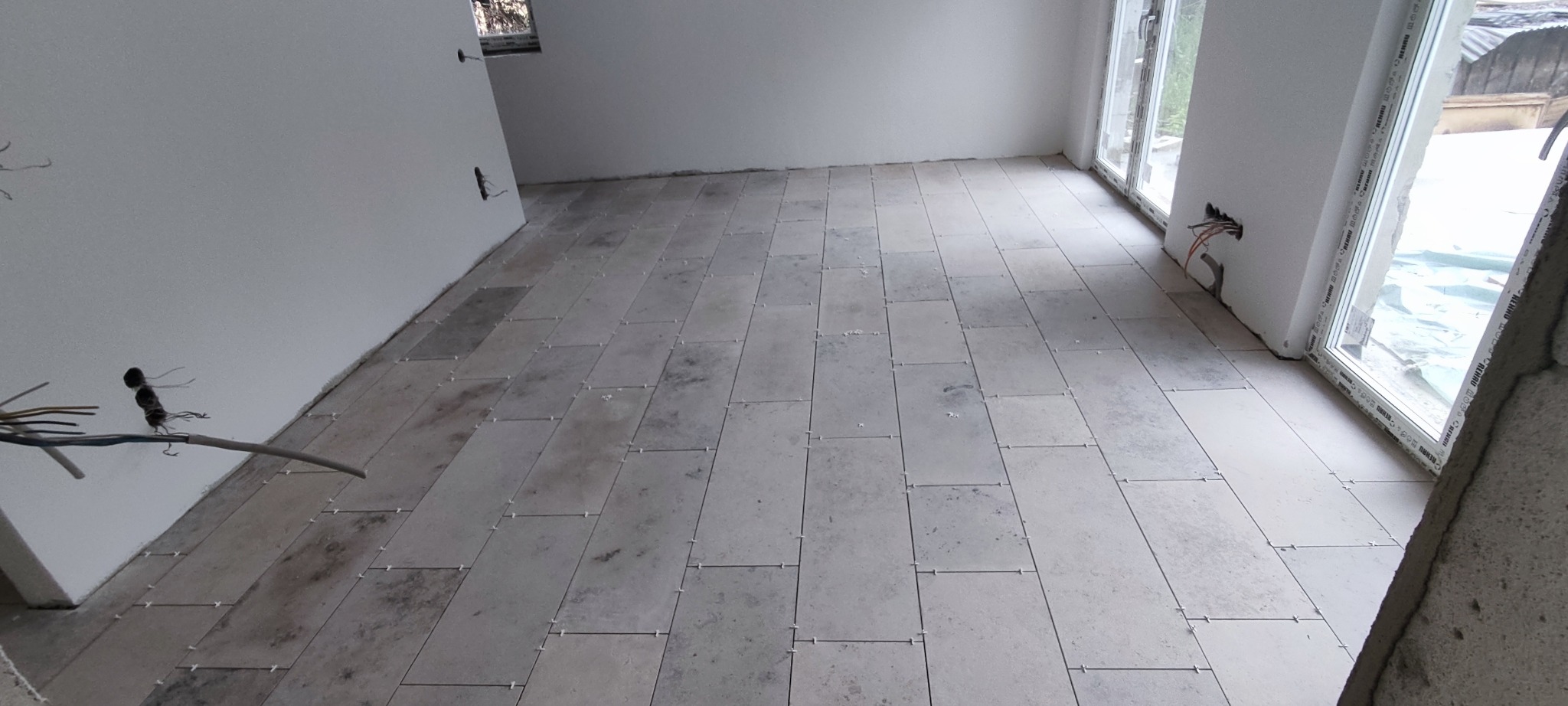 Jasne, prostokątne płytki podłogowe ułożone w nowym pomieszczeniu z oknami. Widoczne krzyżyki dystansowe i wystające kable elektryczne.