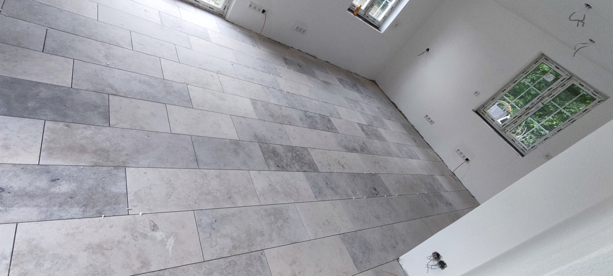 Ułożona podłoga z szarych, prostokątnych płytek w jasnym pomieszczeniu z oknami. Widoczne krzyżyki dystansowe między płytkami. Świeżo położona podłoga.