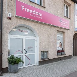 Fasada biura Freedom Nieruchomości z różowym szyldem i wejściem z rośliną w donicy. Widoczny adres na budynku i okno z wizerunkiem kobiety.