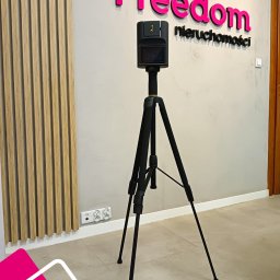 Nieruchomości Sławomir Maliński - Kamera 3D na statywie w biurze Freedom Nieruchomości, gotowa do wirtualnego spaceru. Nowoczesny sprzęt, logo firmy na ścianie, drewniane panele dekoracyjne.