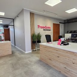 Nowoczesne biuro agencji nieruchomości Freedom z logo na ścianie, biurkami i roślinami. Jasne wnętrze z elementami drewna i szarości.