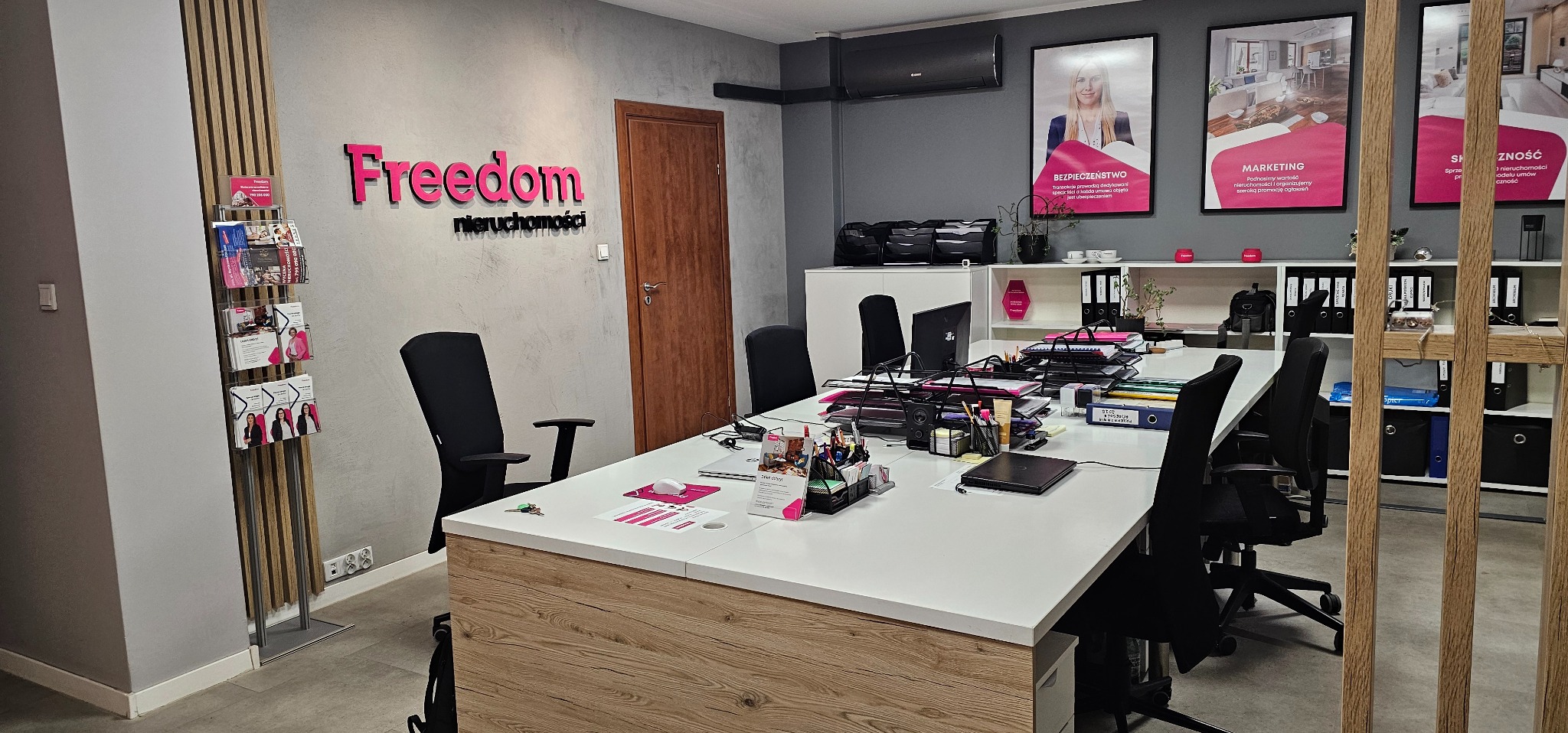 Biuro nieruchomości Freedom: logo na ścianie, plakaty marketingowe, duży stół z dokumentami i krzesłami. Nowoczesne wnętrze z elementami drewna.