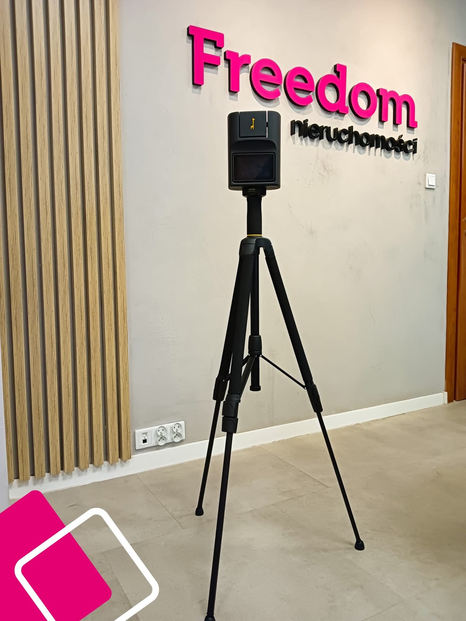 Kamera 3D na statywie w biurze Freedom Nieruchomości, gotowa do wirtualnego spaceru. Nowoczesny sprzęt, logo firmy na ścianie, drewniane panele dekoracyjne.
