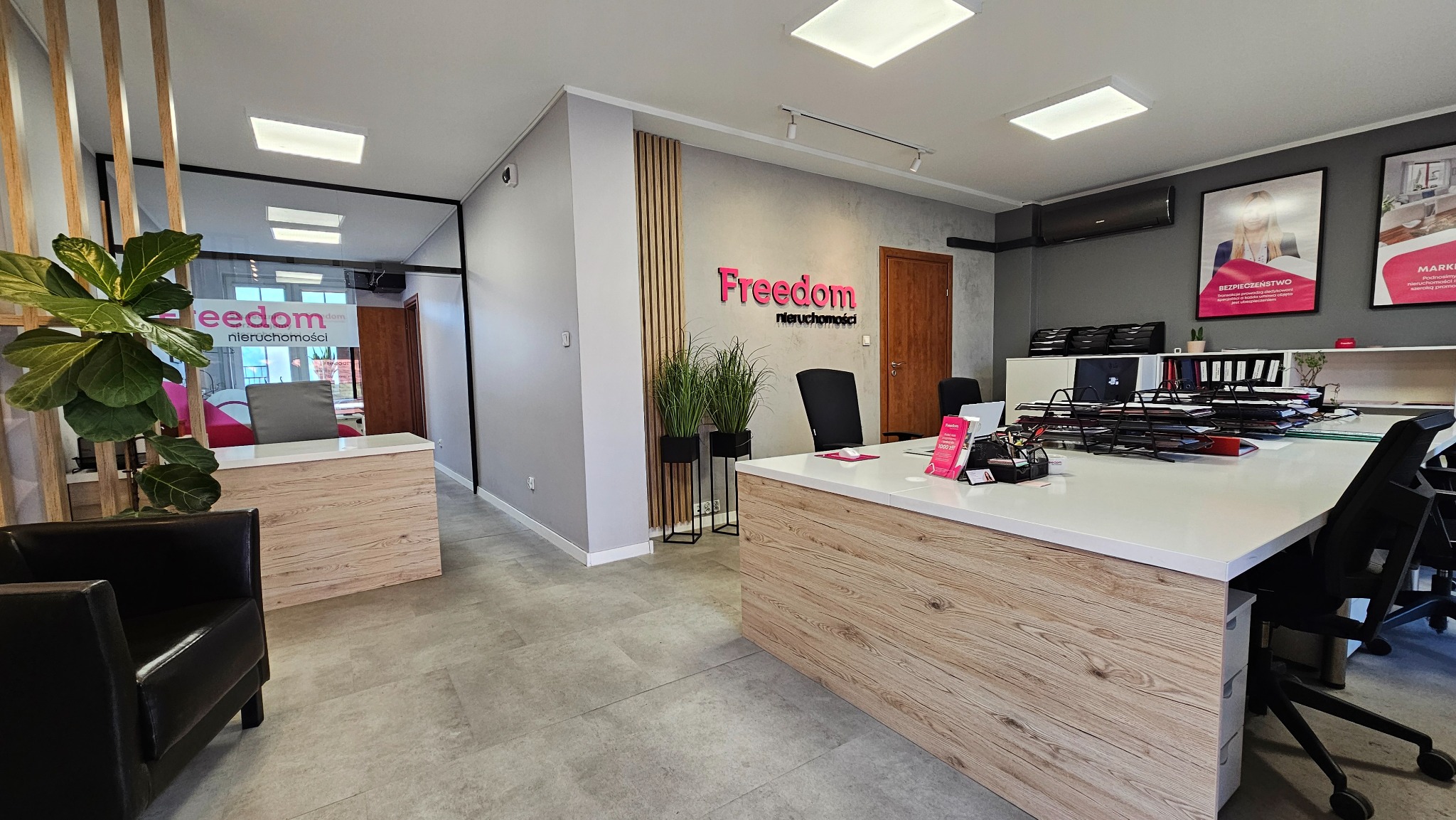 Nowoczesne biuro agencji nieruchomości Freedom z logo na ścianie, biurkami i roślinami. Jasne wnętrze z elementami drewna i szarości.