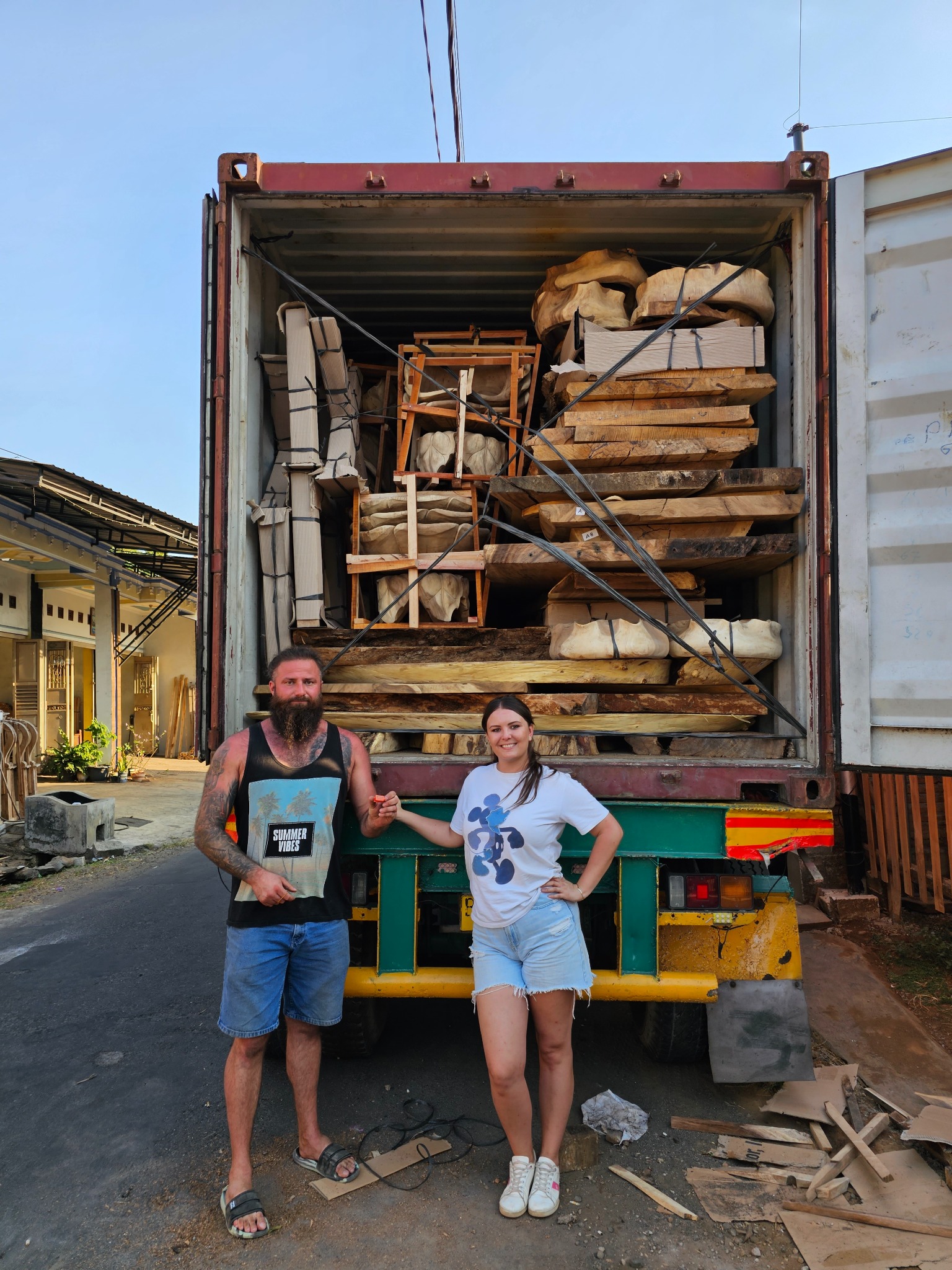 Otwarty kontener transportowy wypełniony drewnem i elementami meblowymi, z parą stojącą przed nim. Widoczne deski, blaty i konstrukcje krzeseł.