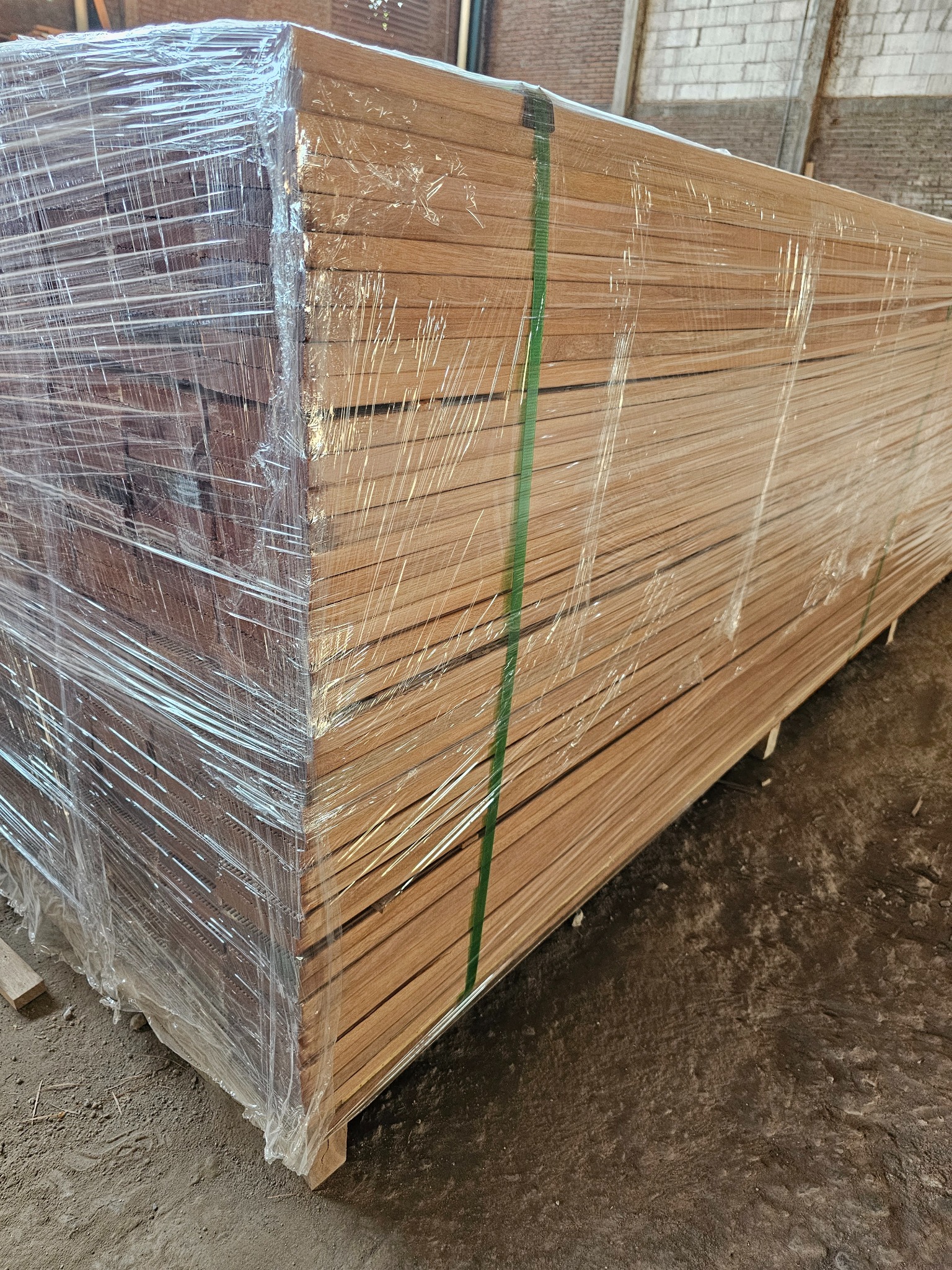 Stos desek tarasowych z jasnego drewna, zabezpieczonych folią i spiętych zielonymi taśmami, składowanych w magazynie. Widoczne ślady magazynowania na podłożu.