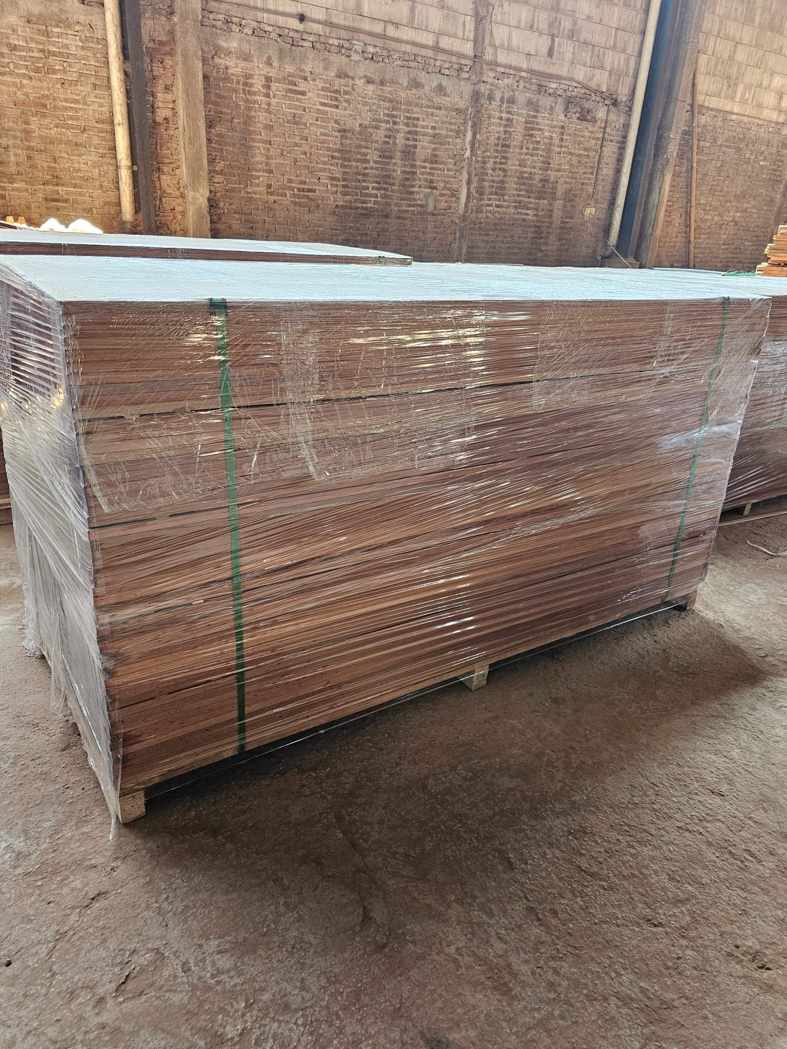 Stos desek tarasowych z egzotycznego drewna, zabezpieczonych folią i spiętych taśmą, na palecie w magazynie z ceglaną ścianą w tle. Widoczne ślady użytkowania.