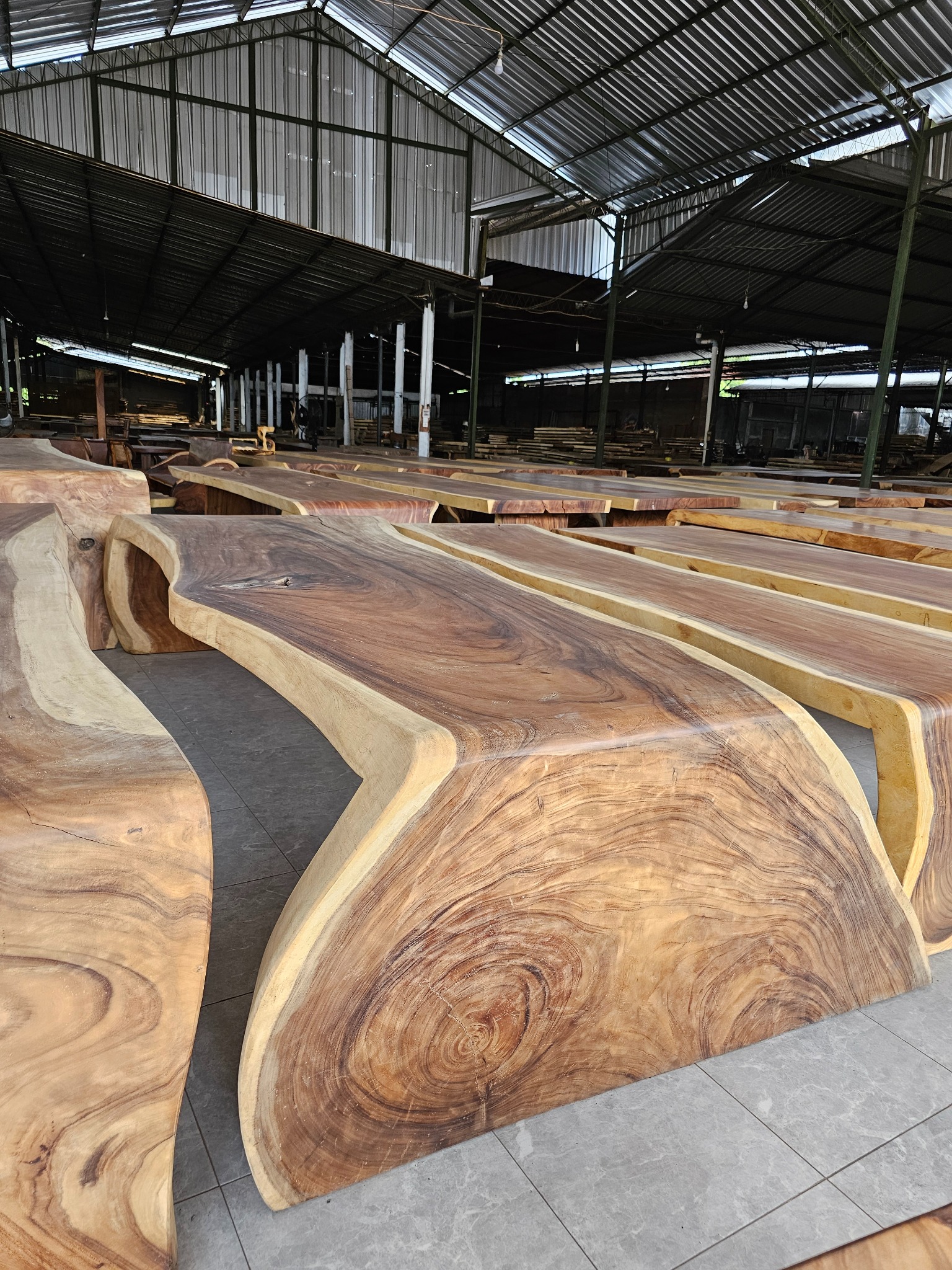 Surowe, drewniane blaty stołowe z widocznym usłojeniem, ułożone rzędami w hali produkcyjnej z metalową konstrukcją dachu. Blaty o nieregularnych kształtach, przygotowane do dalszej obróbki.