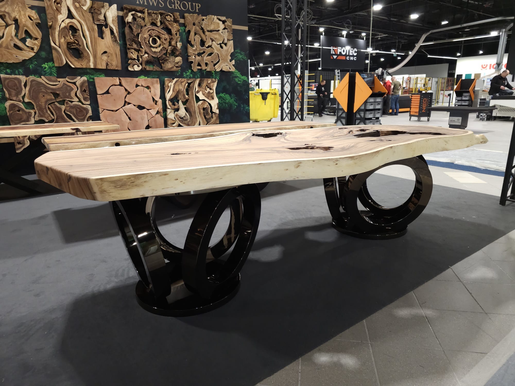 Stół z blatem z litego drewna o nieregularnym kształcie i czarnych, okrągłych nogach, prezentowany na targach. W tle dekoracje ścienne z drewna.