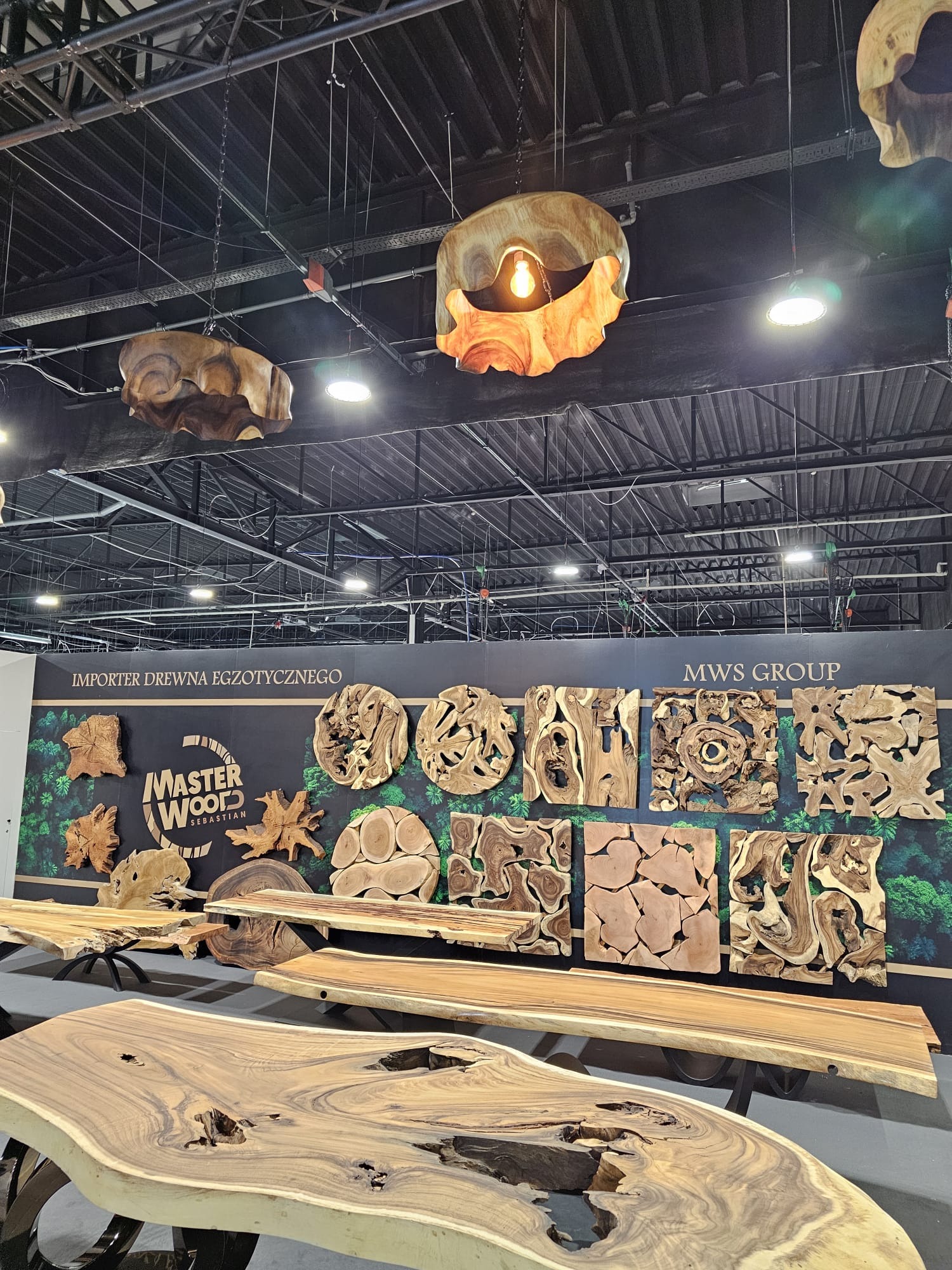 Ekspozycja na targach: blaty stołów z egzotycznego drewna o nieregularnych kształtach, drewniane lampy wiszące i dekoracje ścienne z drewna na tle stoiska Master Wood.