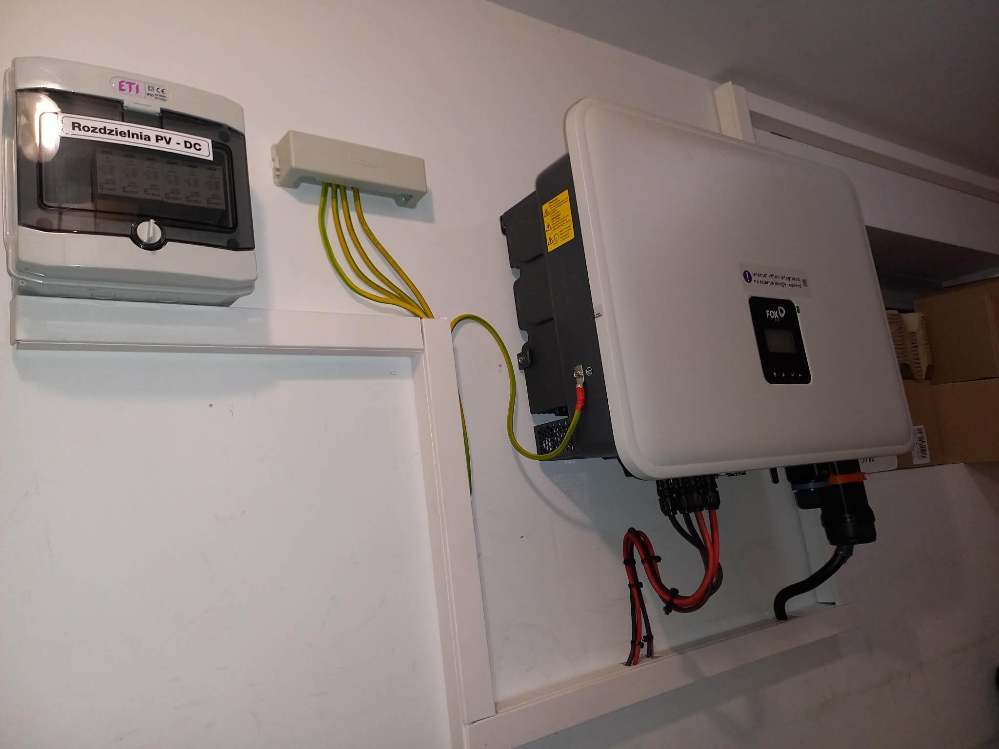 Rozdzielnia PV-DC i falownik na ścianie, widoczne kable. Elementy instalacji fotowoltaicznej wewnątrz budynku, po montażu. Szare i białe obudowy urządzeń.