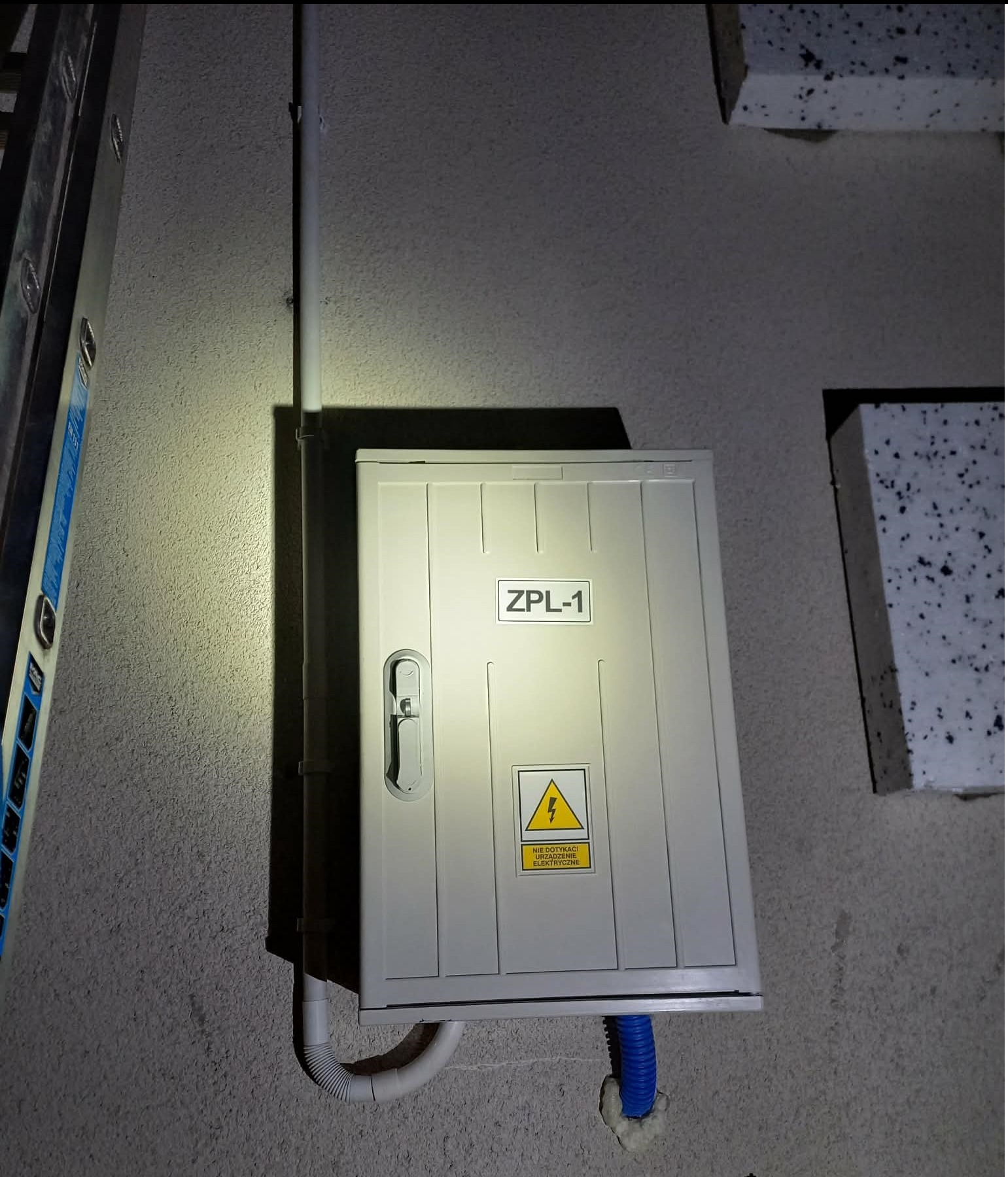 Zamknięta, biała skrzynka elektryczna ZPL-1 z ostrzeżeniem o napięciu, zamontowana na szarej, chropowatej ścianie budynku. Widoczne rury ochronne i fragment drabiny.