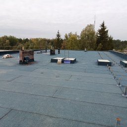 GIT-DACH - Płaski dach pokryty papą termozgrzewalną z kominem i elementami instalacji odgromowej na tle lasu i pochmurnego nieba. Widoczne narzędzia i materiały budowlane.