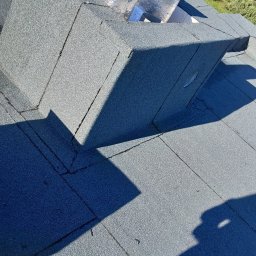 GIT-DACH - Szczegółowe ujęcie płaskiego dachu pokrytego papą z widocznym kominem wentylacyjnym zabezpieczonym folią aluminiową. Ciemne cienie podkreślają strukturę pokrycia.