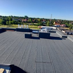 GIT-DACH - Szary, płaski dach pokryty papą termozgrzewalną z kominami wentylacyjnymi i świetlikiem. W tle zabudowa jednorodzinna i zieleń w słoneczny dzień.