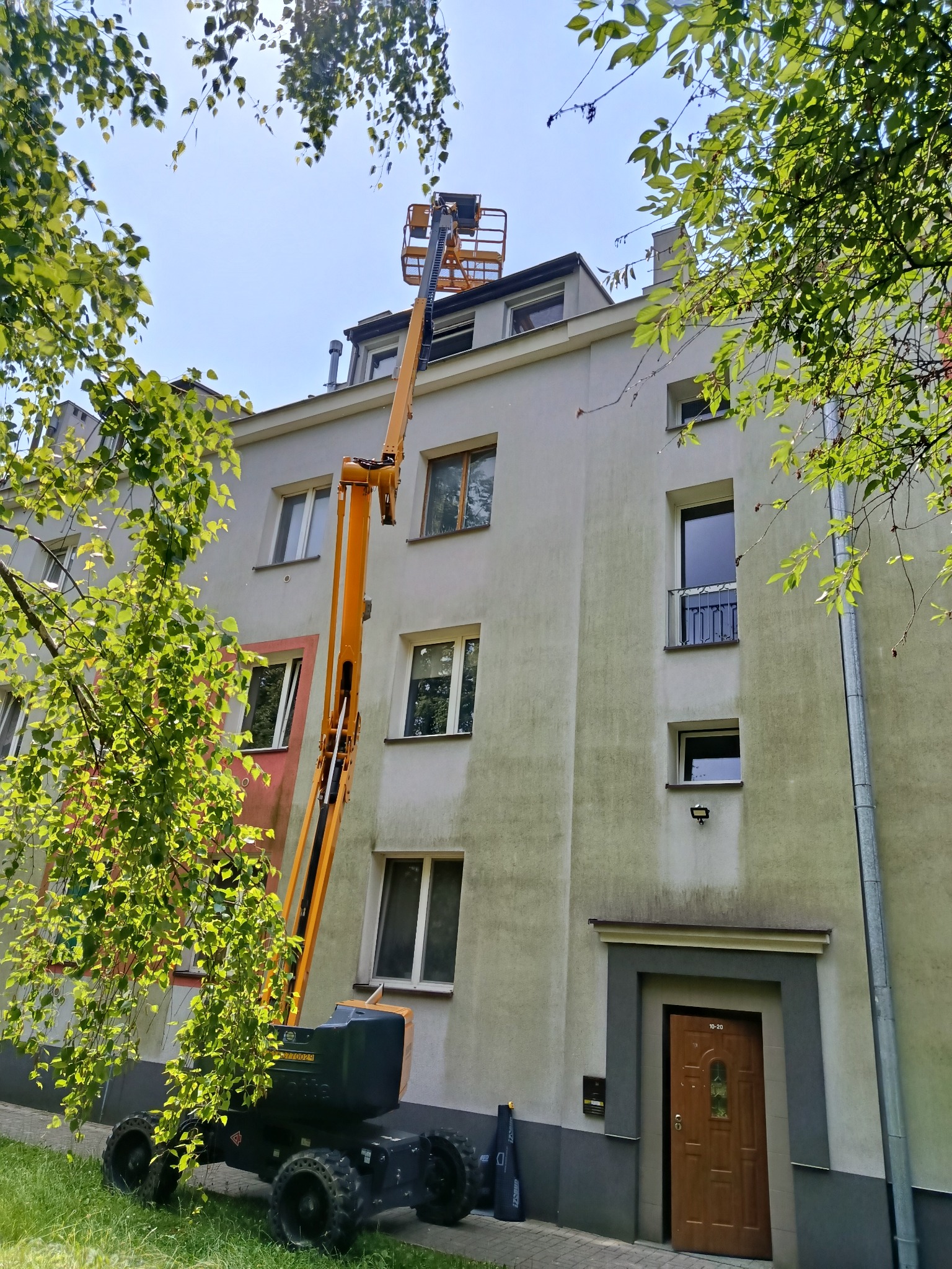 Żółty podnośnik koszowy przy budynku z szarą elewacją, widok z dołu, częściowo zasłonięty przez gałęzie drzew. Prace na wysokościach.