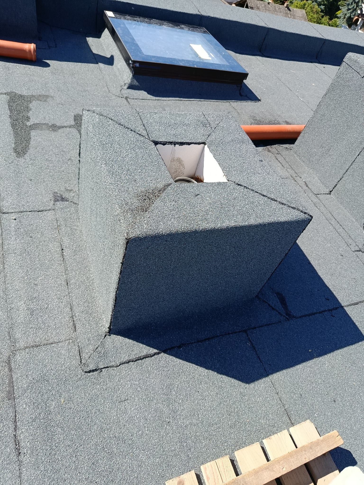 Dach pokryty papą z kominem wentylacyjnym i oknem dachowym. Widoczne detale łączeń i obróbek blacharskich. Fragment palety drewnianej w rogu kadru.