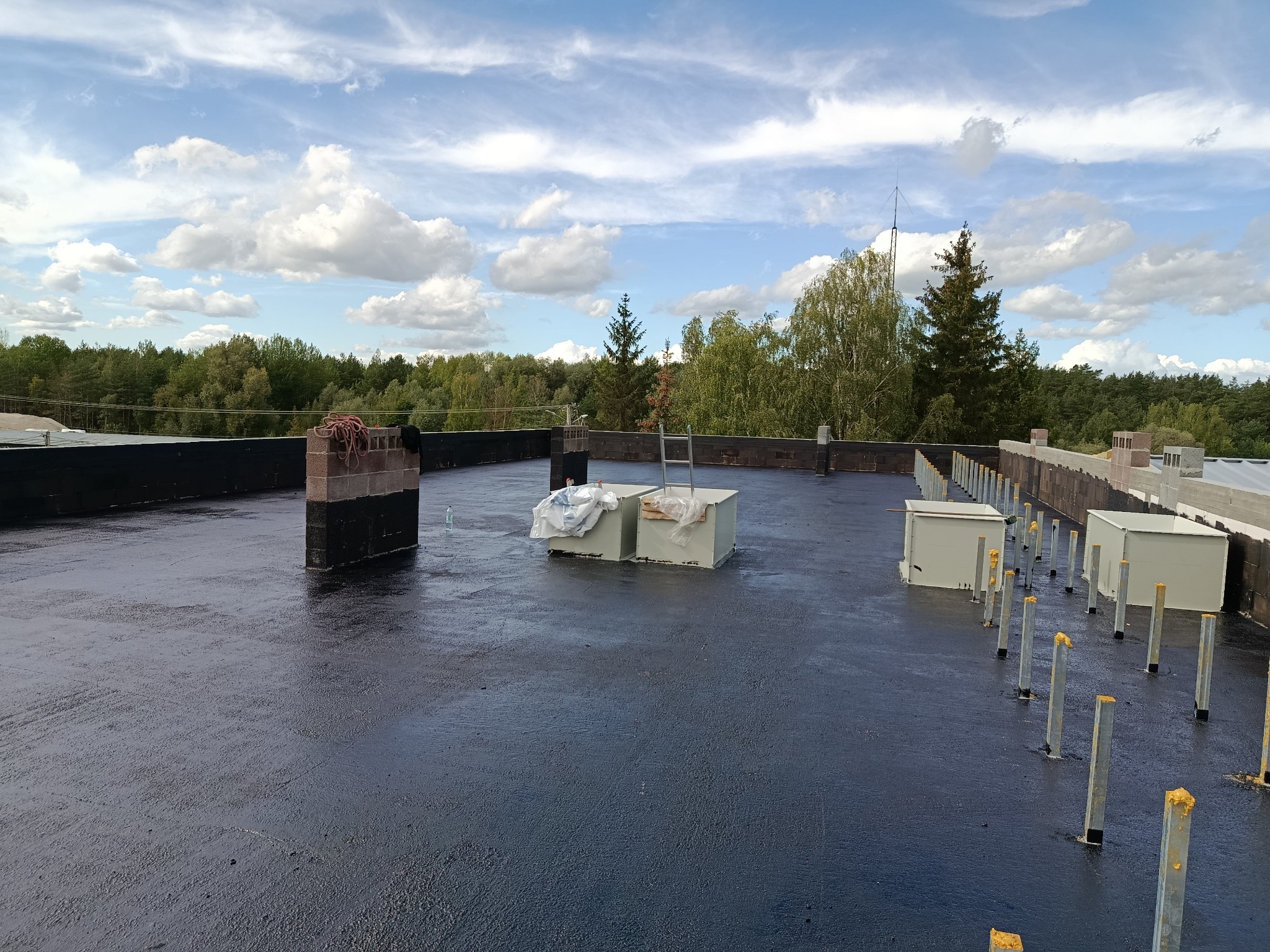 Czarna, płaska powierzchnia dachu z elementami konstrukcyjnymi i widokiem na las w tle, w słoneczny dzień z chmurami.