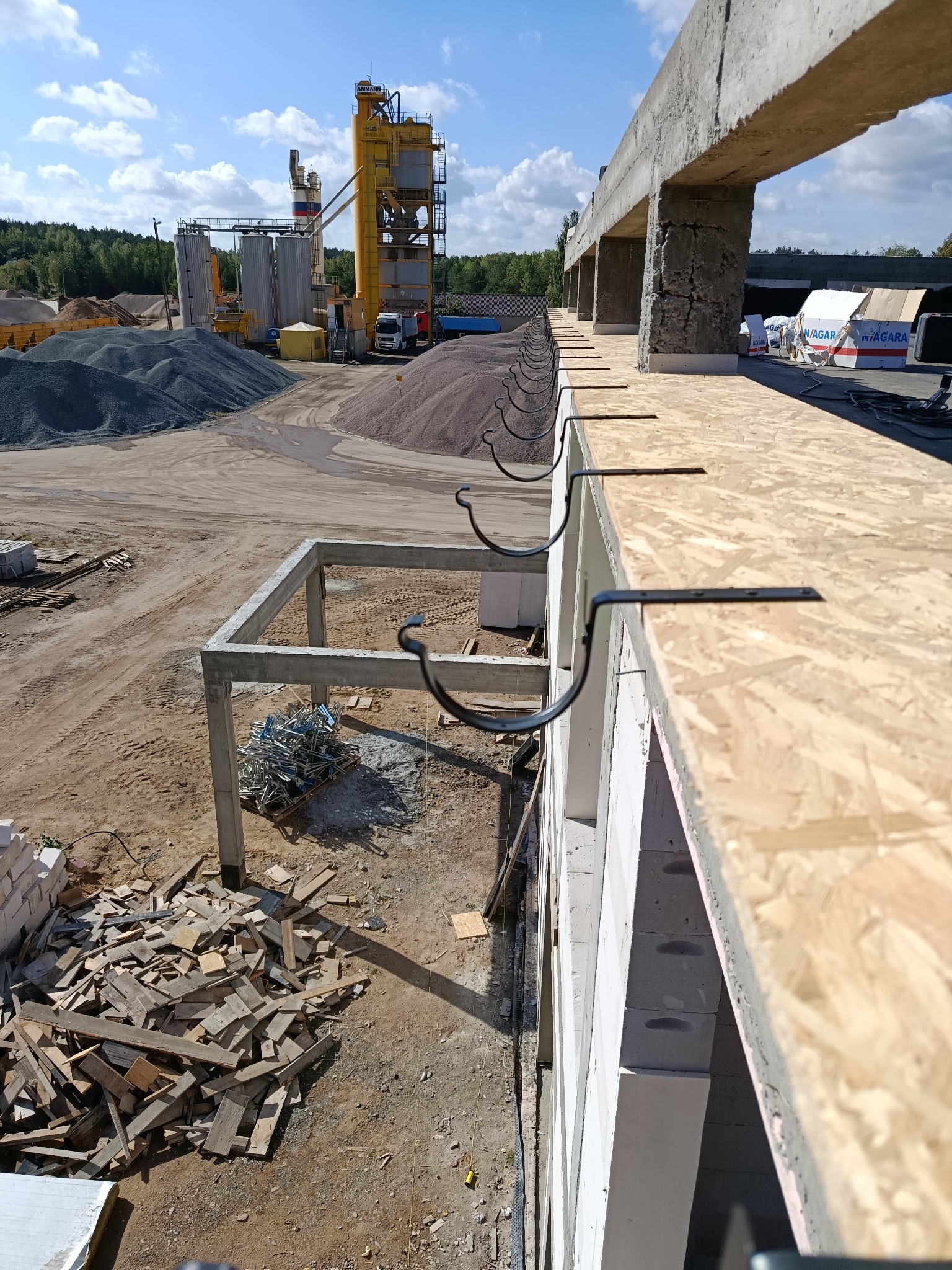 Montaż haków rynnowych na budynku w trakcie budowy. Widoczne elementy konstrukcyjne, płyty OSB i materiały budowlane. W tle żwirownia i błękitne niebo.