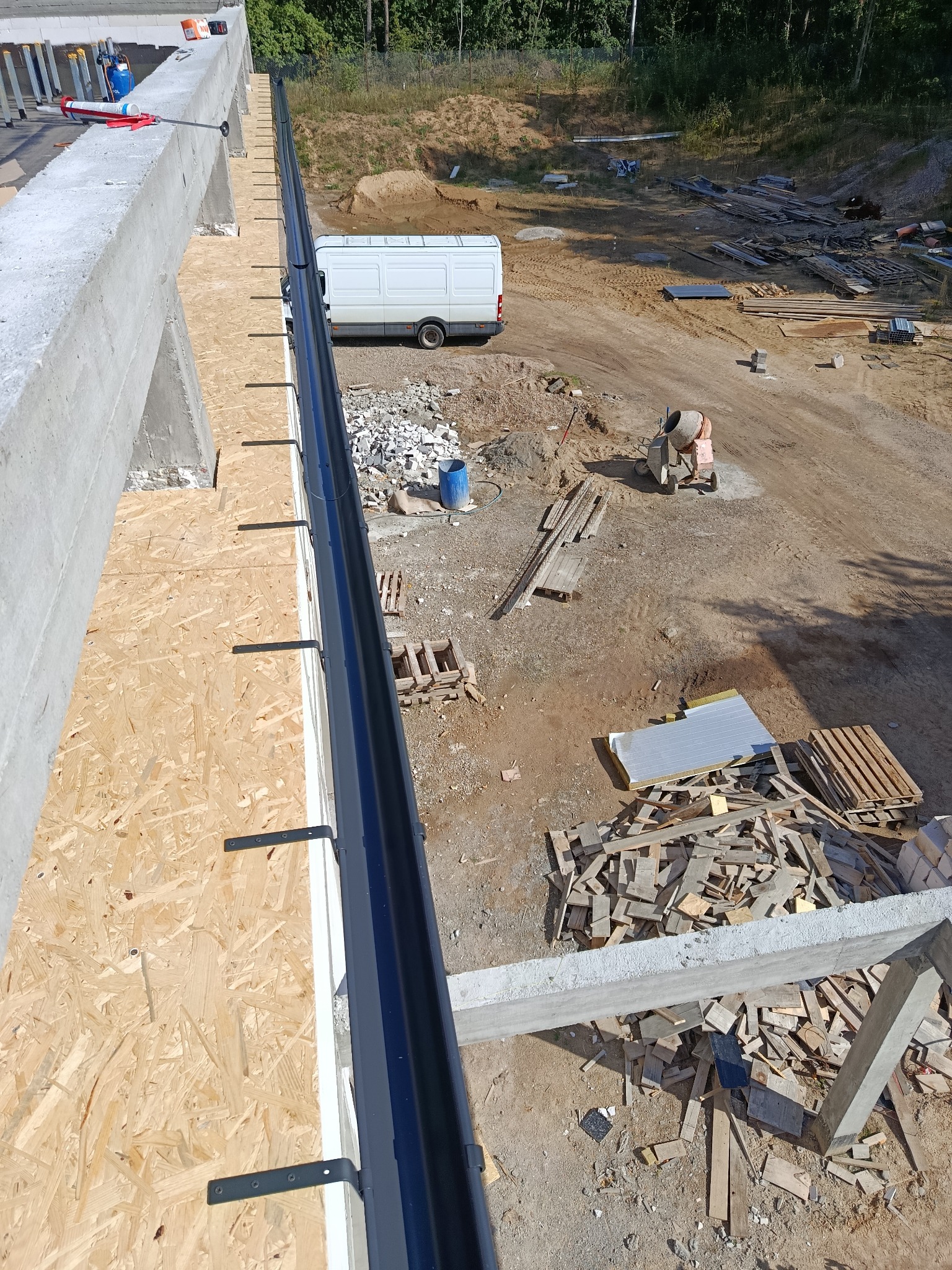 Montaż czarnej rynny dachowej z widocznymi uchwytami na drewnianej płycie OSB, z betonową konstrukcją i placem budowy w tle. Ujęcie z góry.