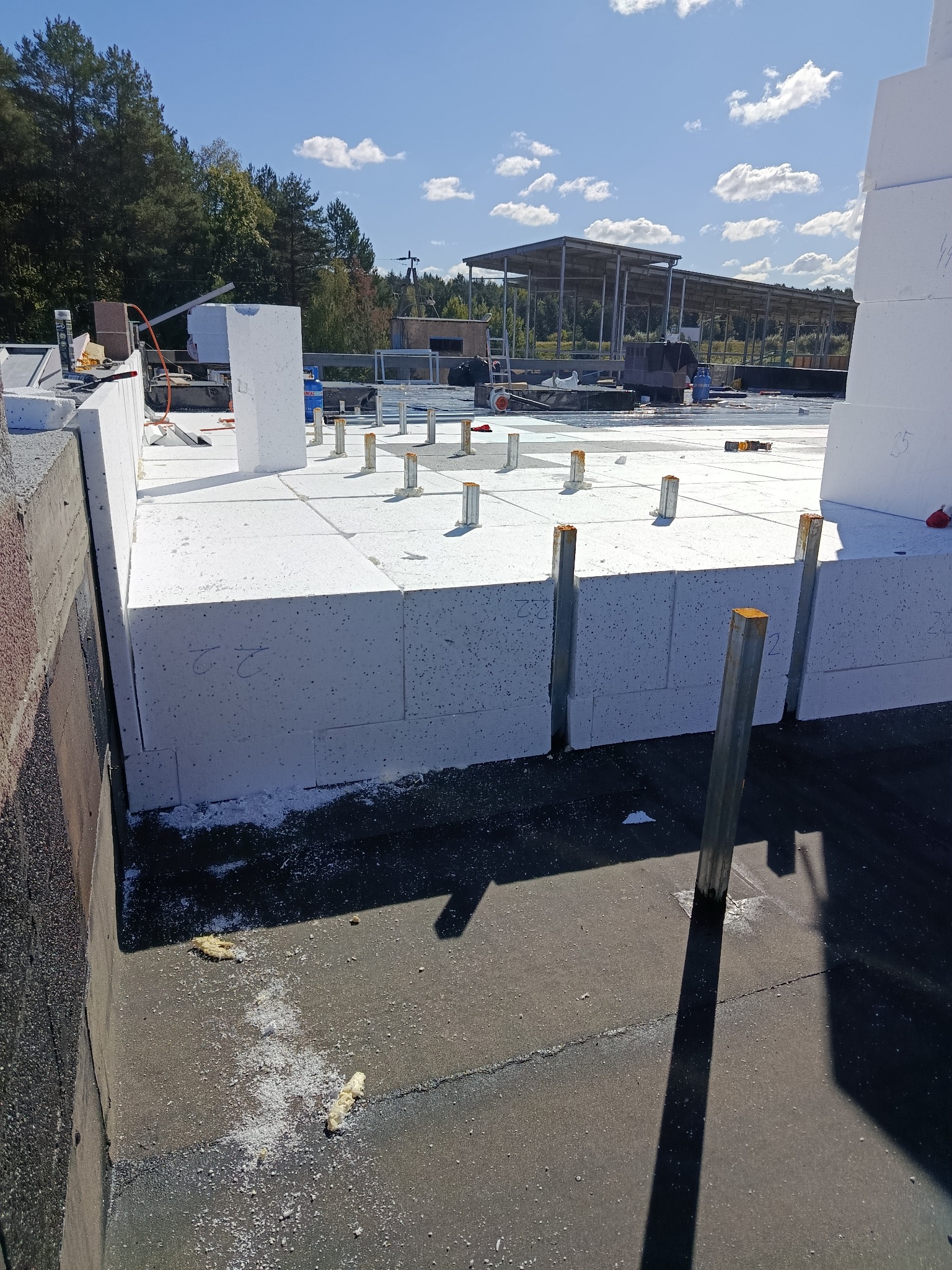 Białe płyty izolacyjne z metalowymi wspornikami na dachu budynku w słoneczny dzień. Widoczne narzędzia i elementy konstrukcyjne. Etap ocieplania dachu.